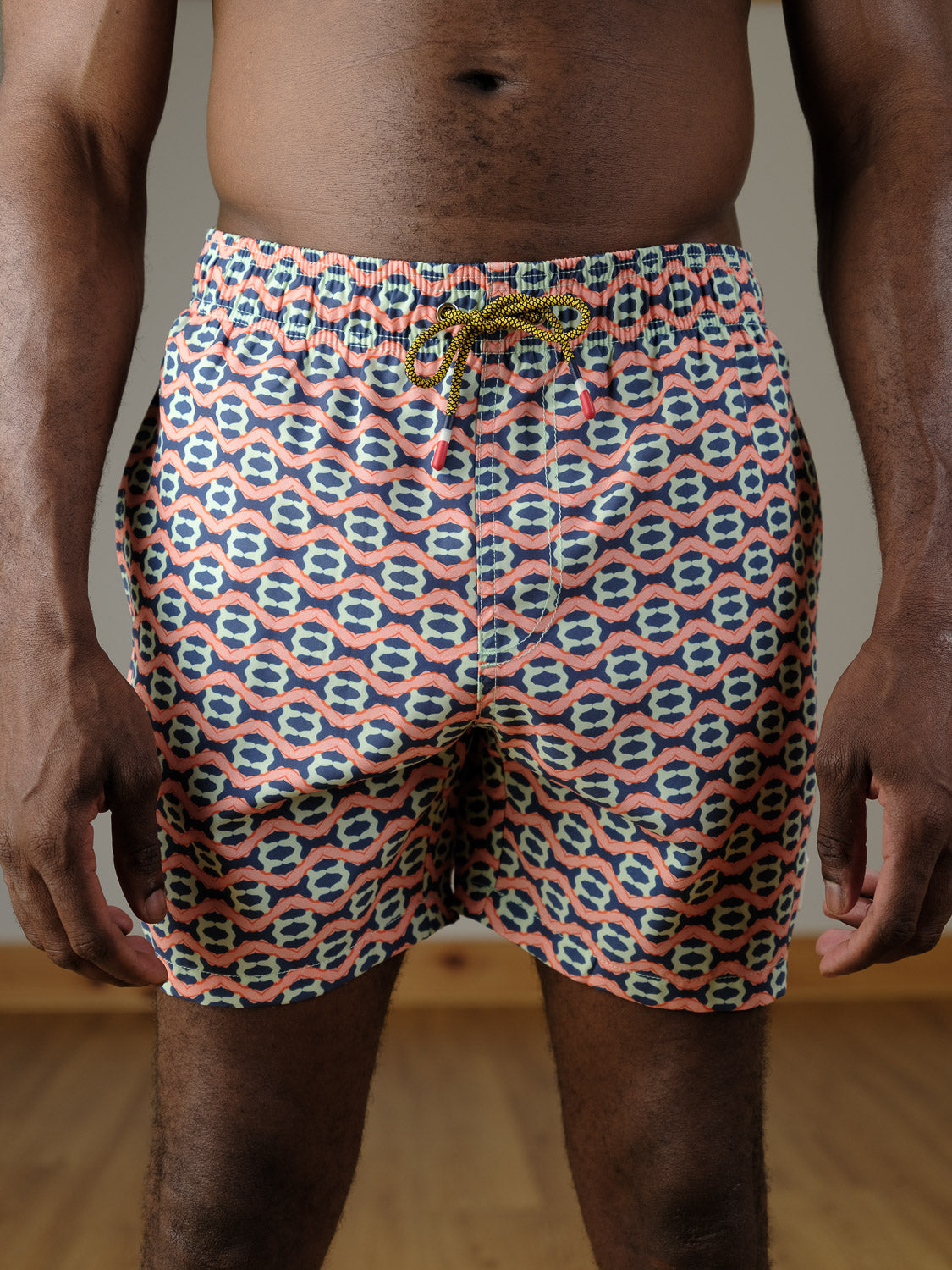 A person indoors on a wooden floor wears ID Menswear's Marbella Fast Dry 4-Way Stretch Swim Trunk, featuring a yellow and black drawstring and an abstract geometric print in pink, blue, and white.