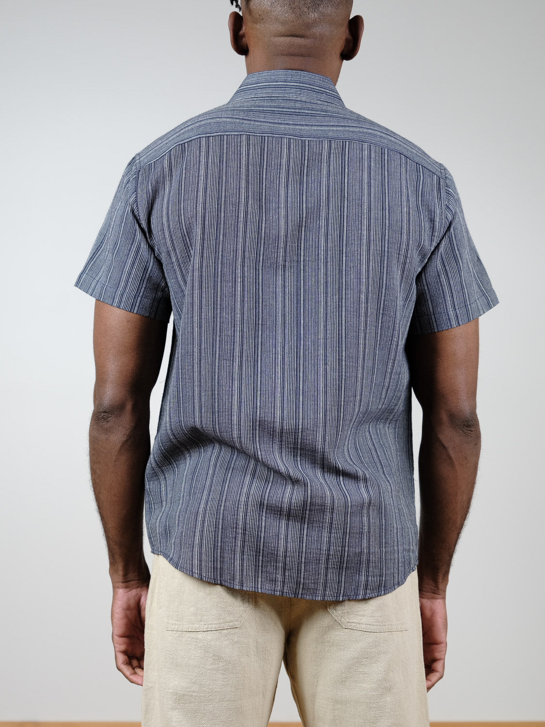 A man faces away from the camera, wearing the ID Menswear Strider Short Sleeve Shirt in blue and white stripes with light beige pants, set against a plain light background.