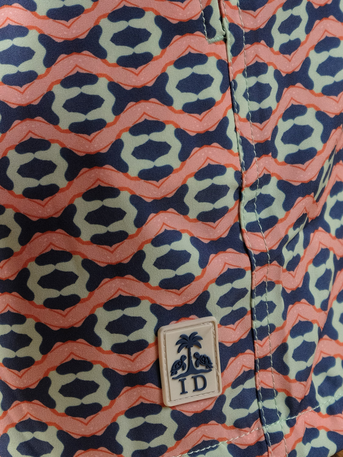 A close-up of Marbella Fast Dry 4-Way Stretch Swim Trunk fabric by ID Menswear, featuring an abstract coral, black, and light green geometric pattern and a small patch with palm trees, turtles, and the letters ID.