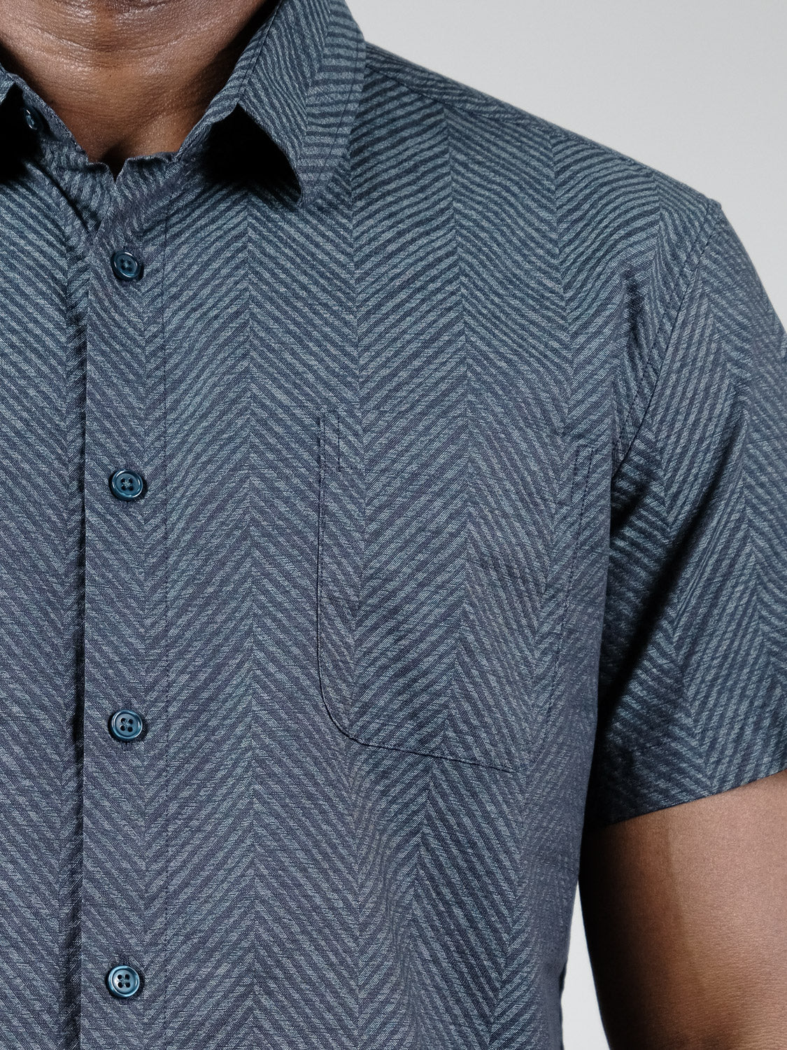 A close-up of a person wearing the Reveller Short Sleeve Shirt by ID Menswear, featuring a relaxed fit, subtle chevron weave, black buttons, collar, and small chest pocket. Only the upper torso and part of the left arm are visible.