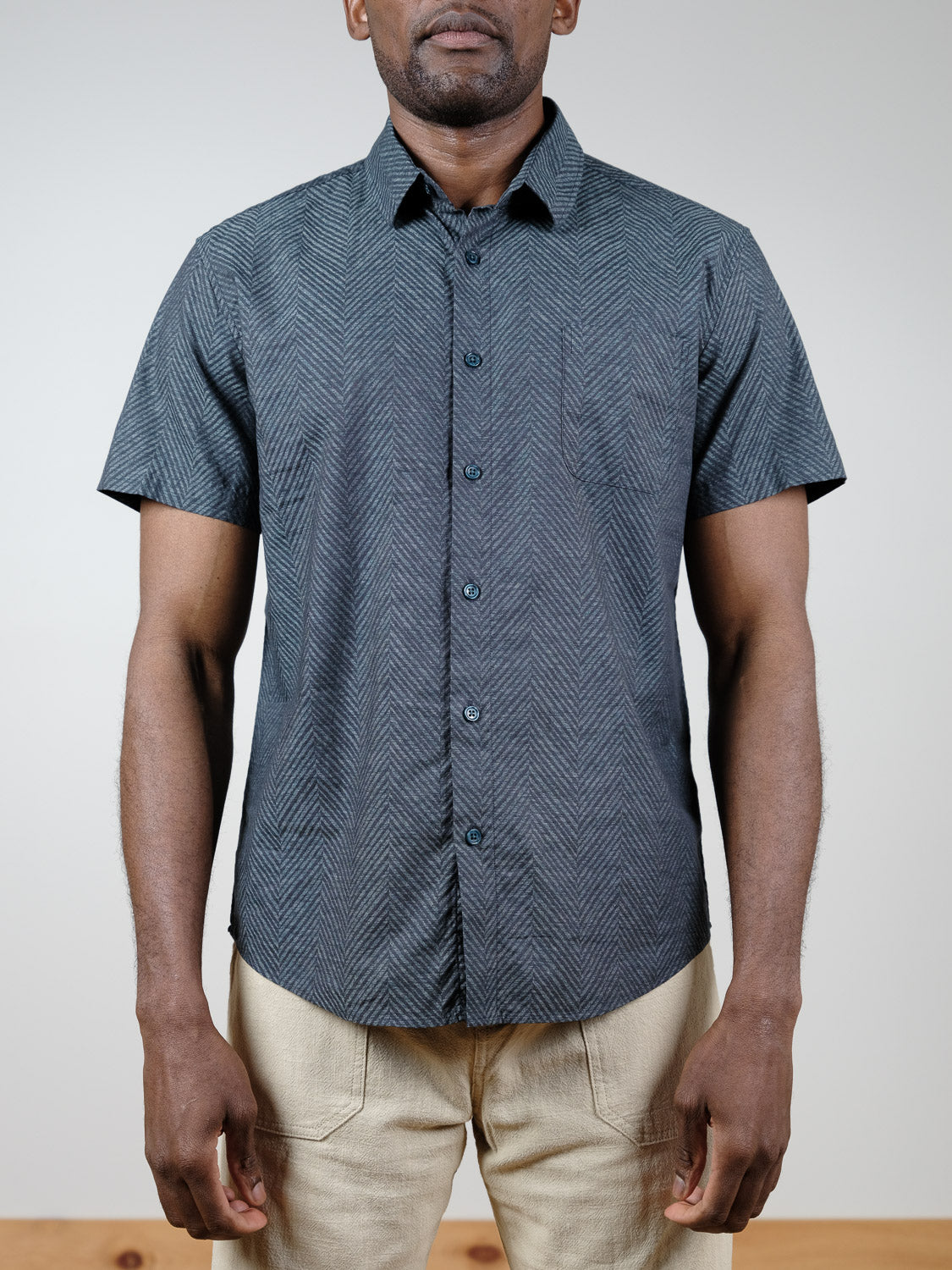 A man wears the Reveller Short Sleeve Shirt by ID Menswear, featuring a relaxed fit and dark geometric weave, paired with light pants against a plain, light background.