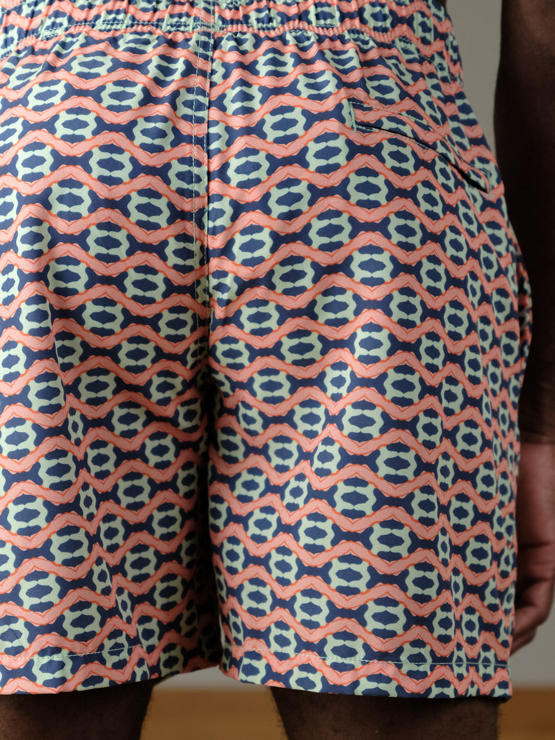 Close-up of a person wearing ID Menswear's Marbella Fast Dry 4-Way Stretch Swim Trunk, featuring a colorful geometric pattern in blue, pink, and beige with repeating ovals and diamonds, an elastic waistband, and a back pocket.