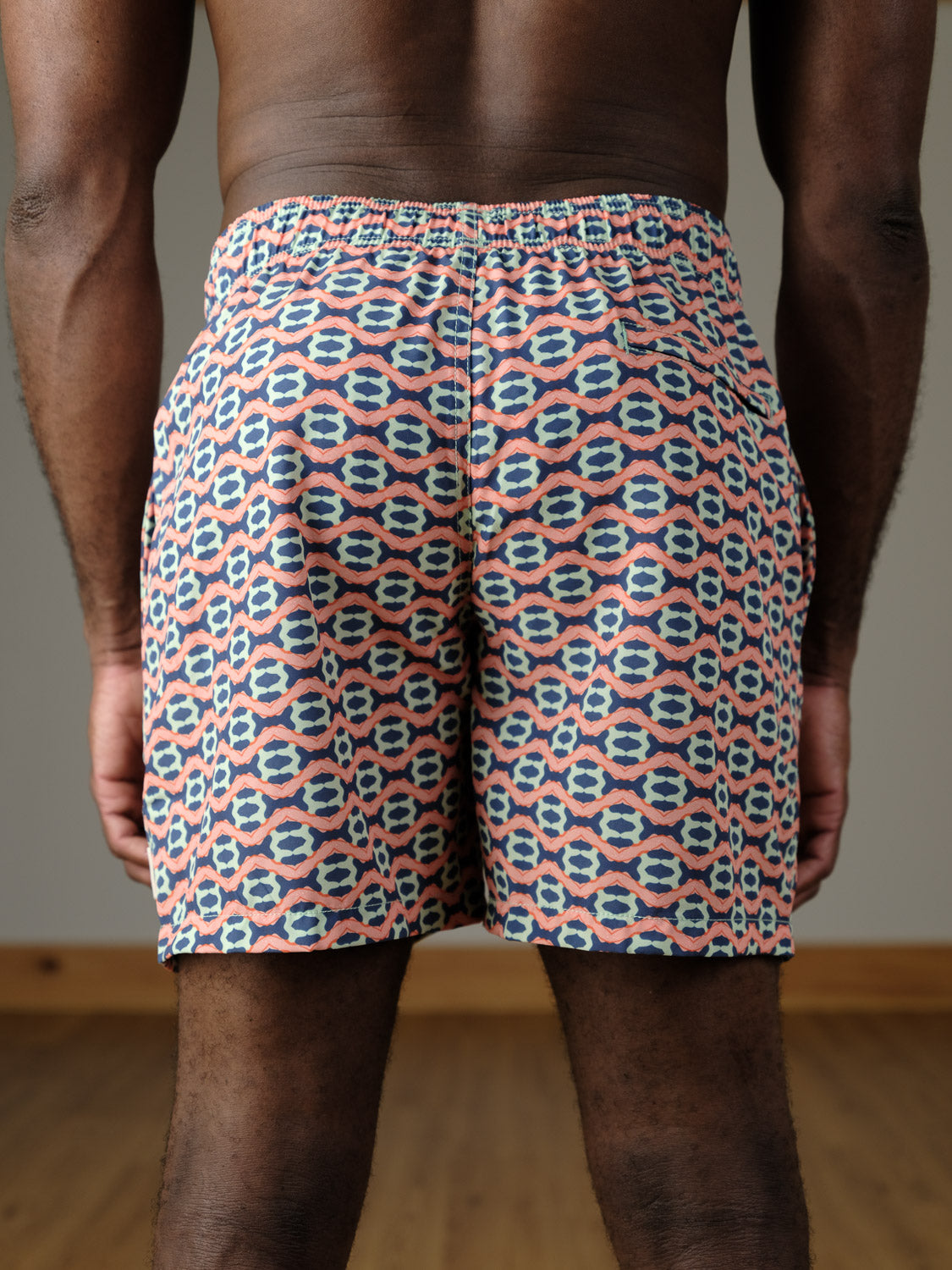 A person faces away, wearing the Marbella Fast Dry 4-Way Stretch Swim Trunk by ID Menswear, featuring a colorful blue, white, and pink geometric design. The background is neutral with a wooden floor.