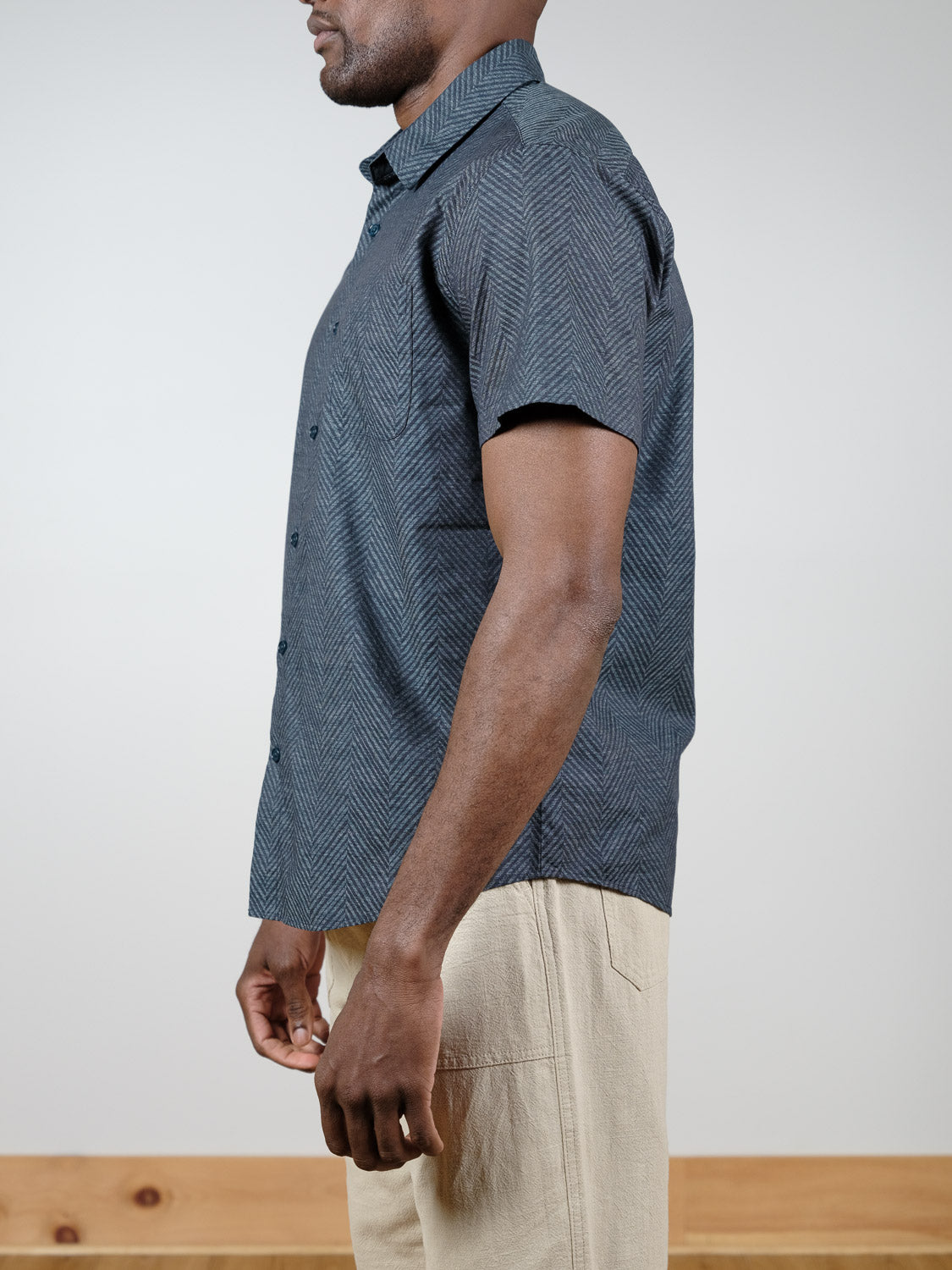 A man stands sideways against a plain background, wearing the ID Menswear Reveller Short Sleeve Shirt—a relaxed fit cotton shirt with a dark geometric weave—paired with light beige pants. Only his upper body is visible, his face partially out of frame.