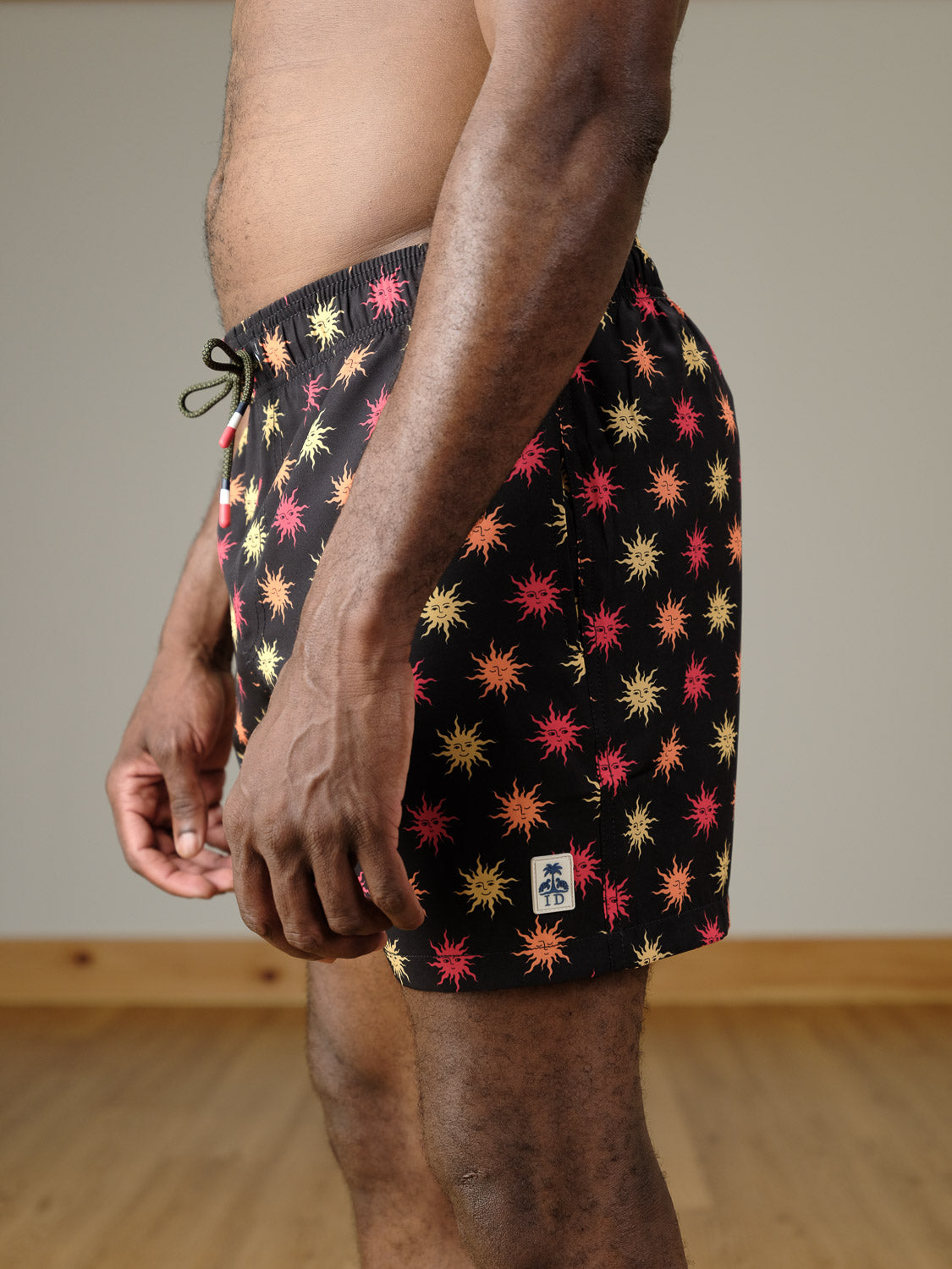 A person wears the Sunshine Fast Dry 4-Way Stretch Swim Trunk by ID Menswear, featuring a black base with a red and yellow starburst pattern, shown indoors from the side and waist down on a wooden floor.
