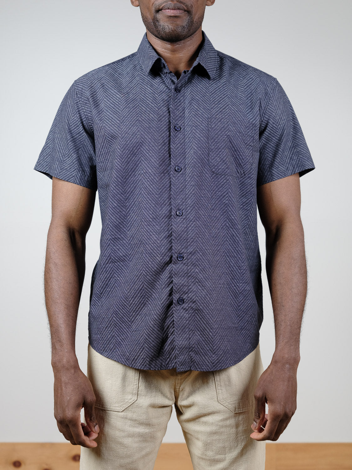 A man wears the ID Menswear Reveller Short Sleeve Shirt in dark blue slub cotton with a subtle chevron pattern, paired with light beige pants, standing against a plain background.