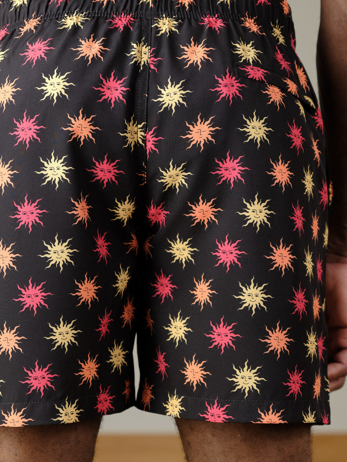Close-up of a person wearing the ID Menswear Sunshine Fast Dry 4-Way Stretch Swim Trunk, showcasing a vibrant yellow, orange, and pink sunburst pattern and an elastic waistband. Only the lower back and upper legs are visible.