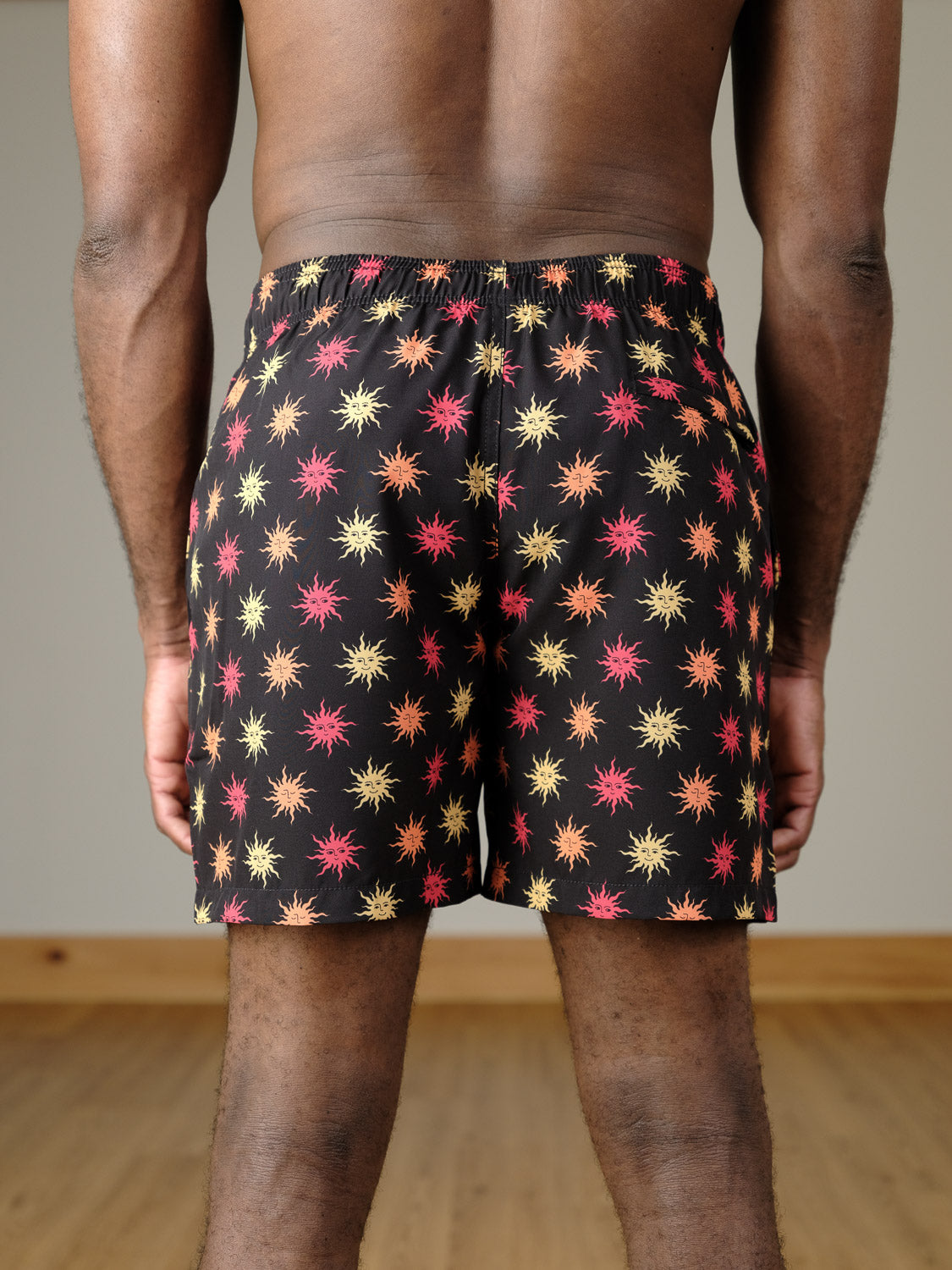 A person stands indoors on a wooden floor, back to camera, wearing ID Menswear's Sunshine Fast Dry 4-Way Stretch Swim Trunk in black with a yellow and pink starburst pattern. The background is plain.