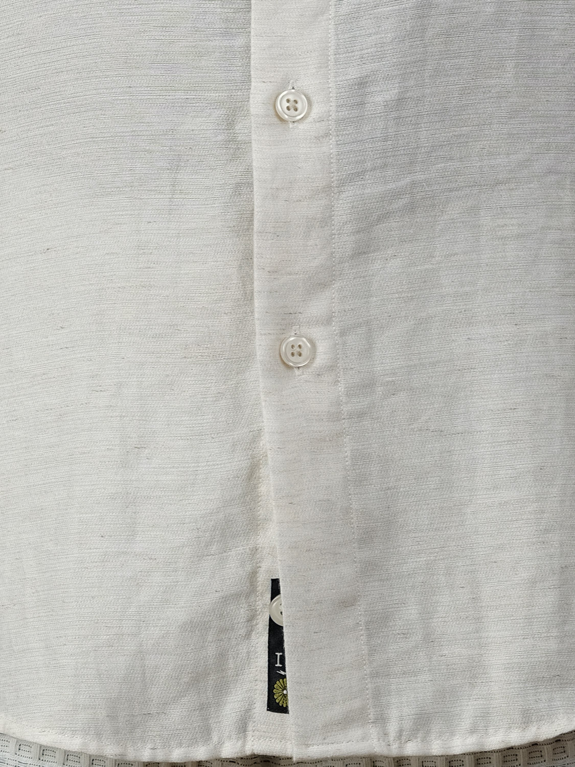 Close-up of the Hiker Short Sleeve Shirt by ID Menswear in relaxed white textured fabric, featuring three visible buttons and a black tag at the hem—an essential addition to your summer basics.