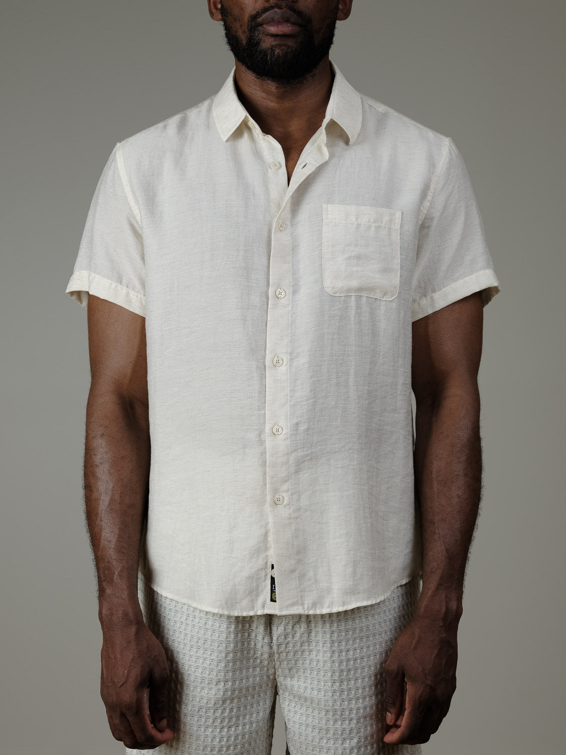 A man wearing the Hiker Short Sleeve Shirt by ID Menswear, a cream linen relaxed-fit top with chest pocket, paired with light-textured pants—an effortless summer basics look against a plain backdrop.