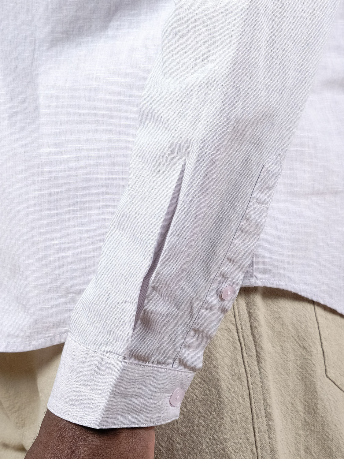 A close-up of a person’s arm wearing the ID Menswear Darwin Long Sleeve Shirt, a light-colored, relaxed fit with visible sleeve cuff, paired with beige textured pants. The lightweight cotton shirt flatters medium to dark skin tones.