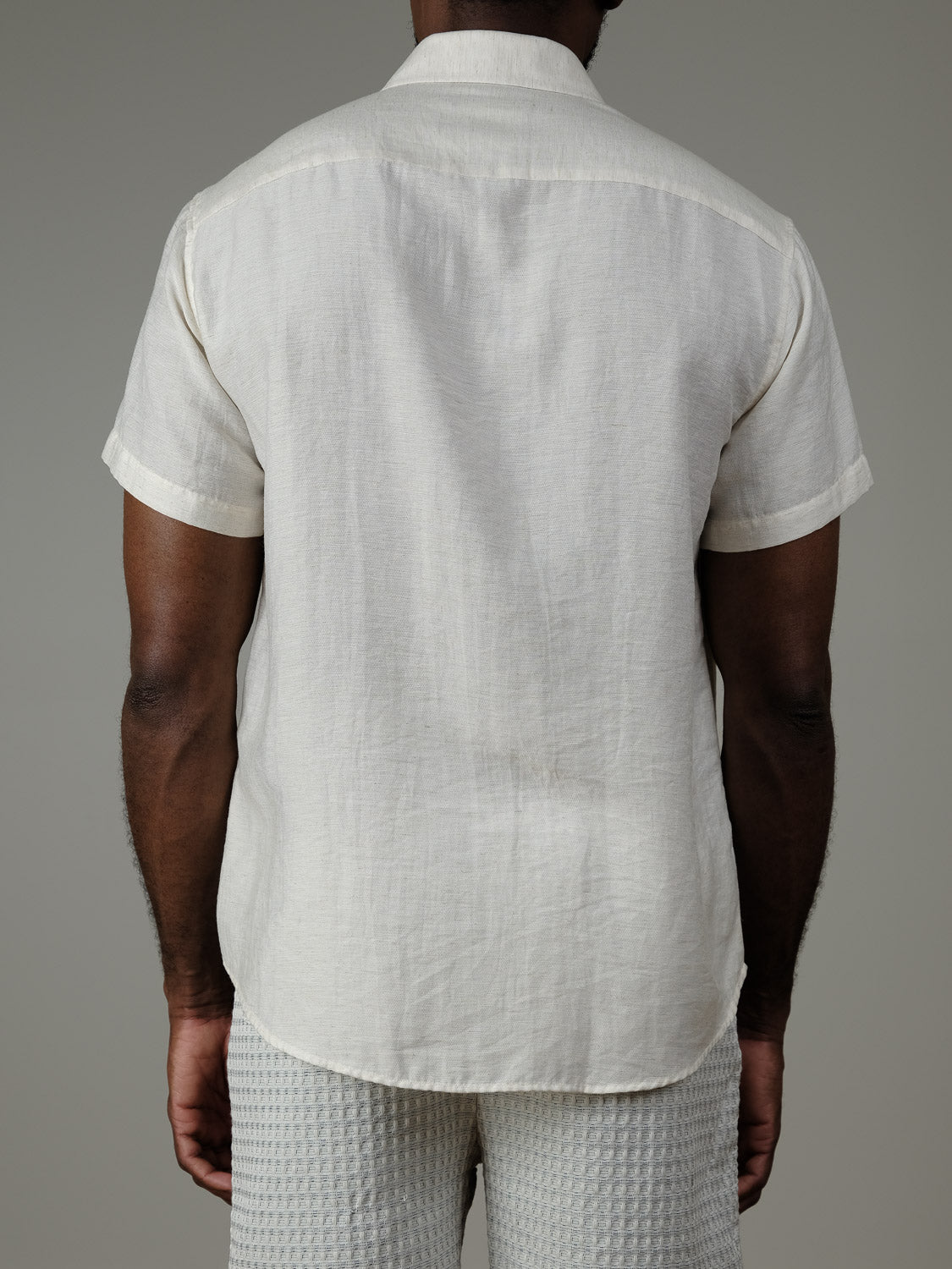 A person is shown from behind wearing the Hiker Short Sleeve Shirt by ID Menswear paired with textured off-white shorts—timeless summer basics—set against a neutral background.