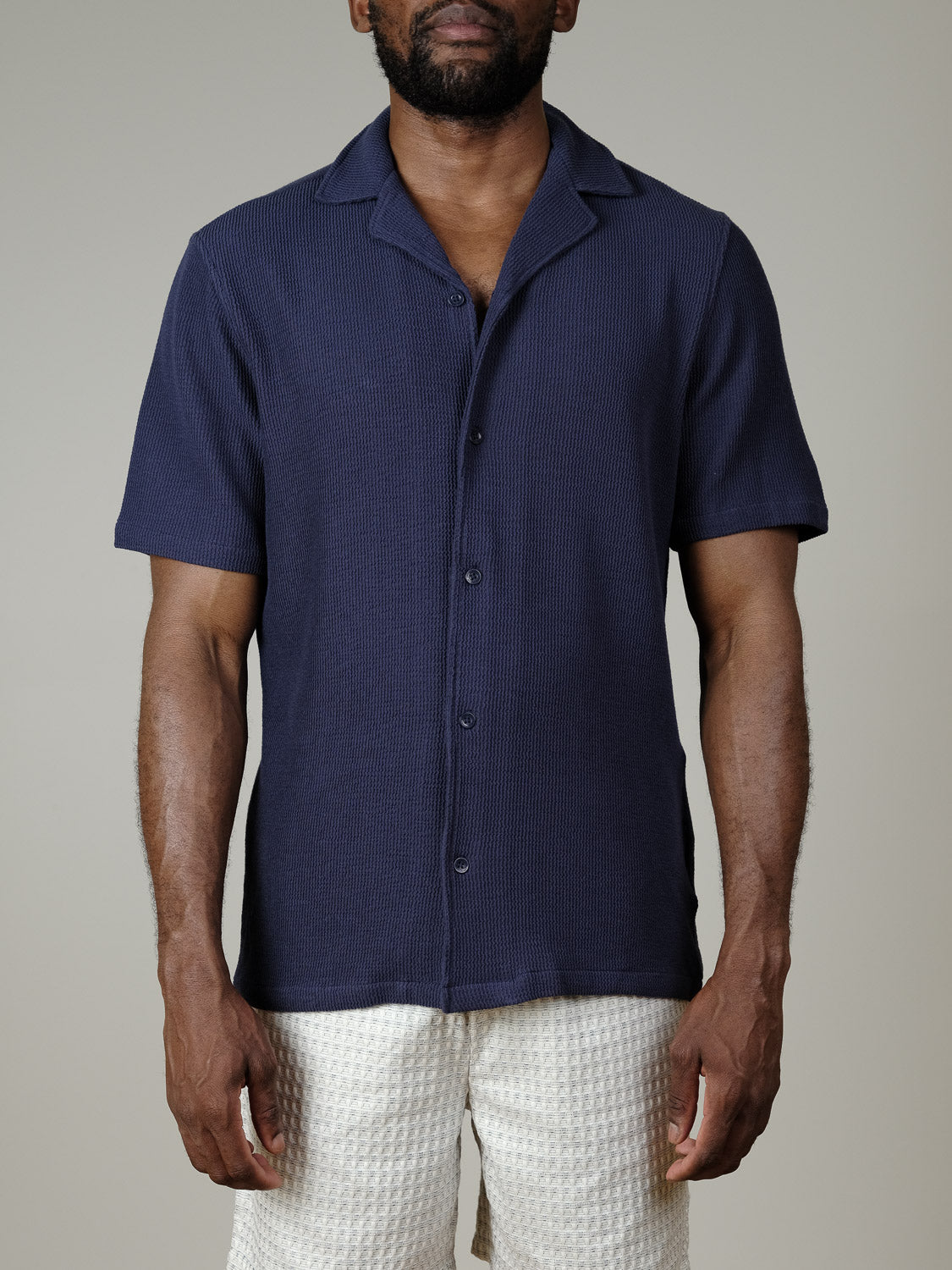 A man stands against a plain light gray background wearing the Casual Friday Archer SS RC Knitted Shirt in dark blue and off-white textured shorts. His face is partially visible.
