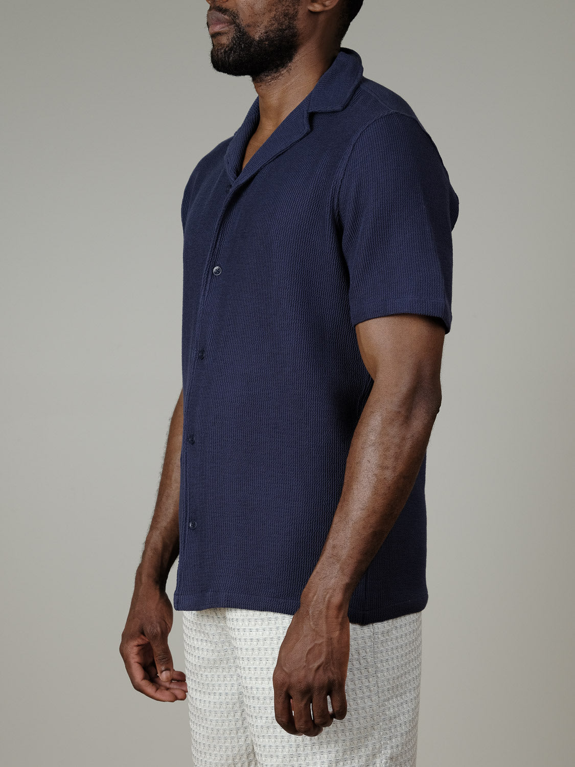A man wears the Archer SS RC Knitted Shirt by Casual Friday in dark blue, paired with white textured shorts, standing against a plain light gray background, facing slightly left with his arms relaxed at his sides.