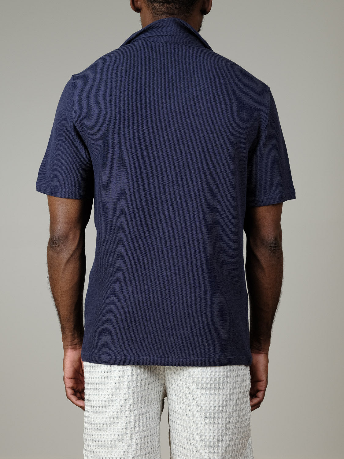A person is shown from the back wearing the Casual Friday Archer SS RC Knitted Shirt in navy and white textured shorts, standing against a plain light gray background.