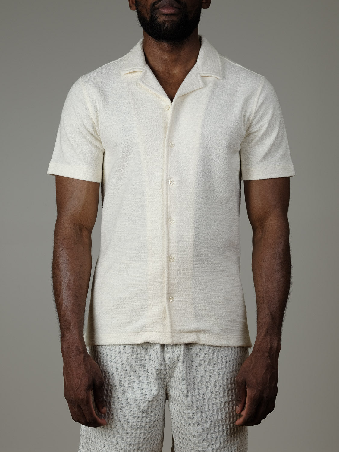 A man stands against a plain gray background, arms at his sides, wearing the Casual Friday Egebjerg Resort Shirt and light textured shorts.
