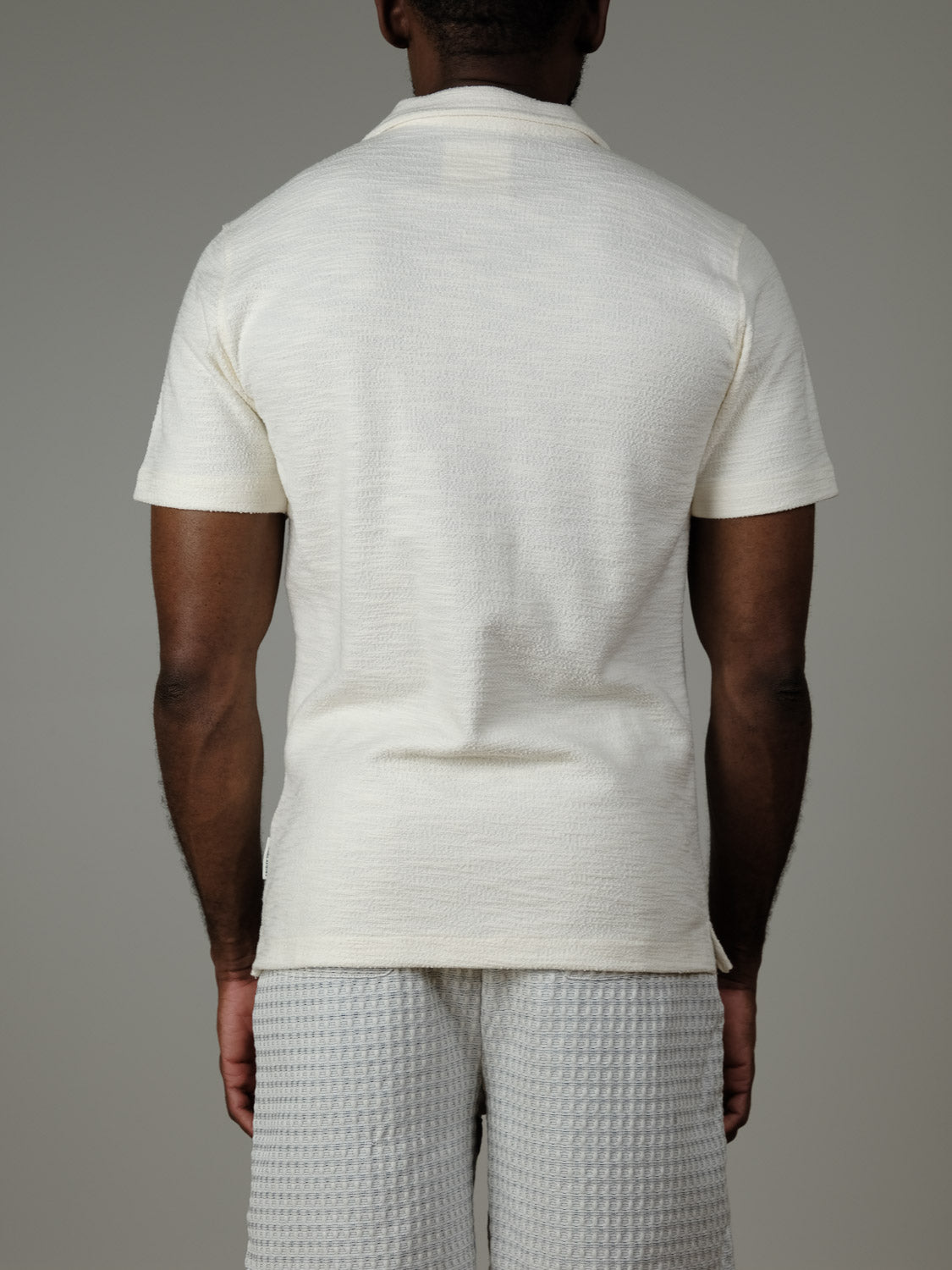 A person faces away from the camera, wearing the Egebjerg Resort Shirt by Casual Friday in off-white textured fabric and light gray shorts—an ideal vacation look—set against a plain gray background.