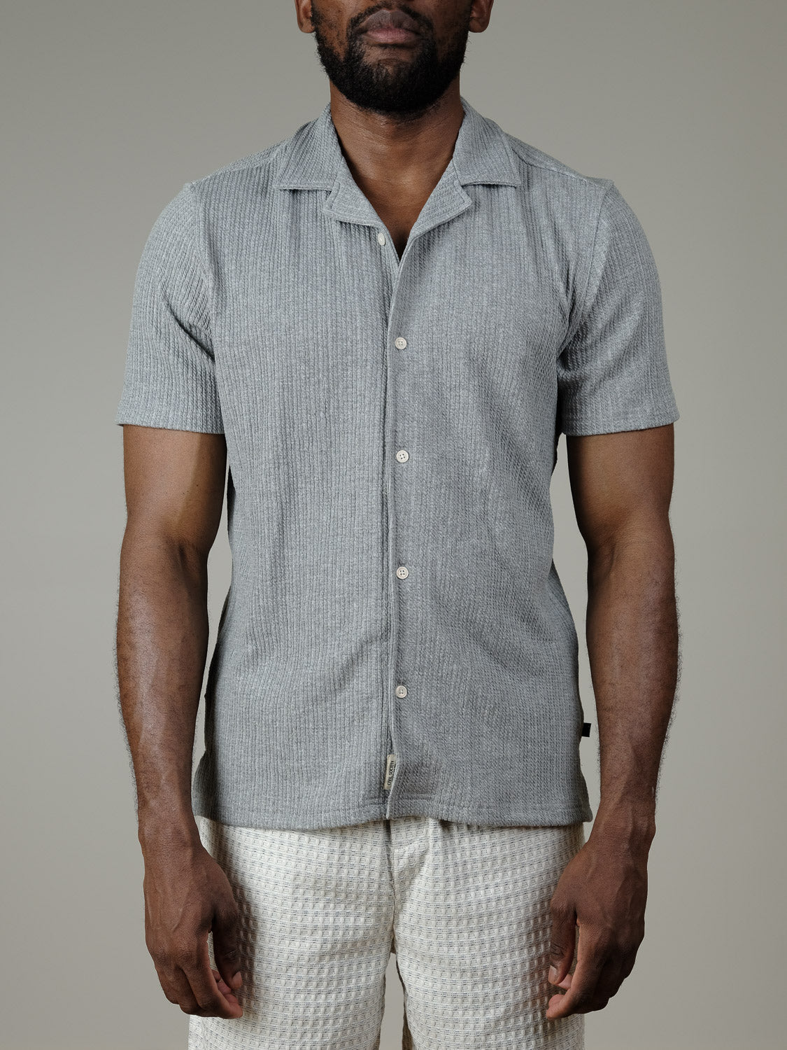 A man stands against a plain background wearing the Civil Society Viggo Knit Resort Shirt in light grey and textured white shorts. The image is cropped at the neck and above the knees.