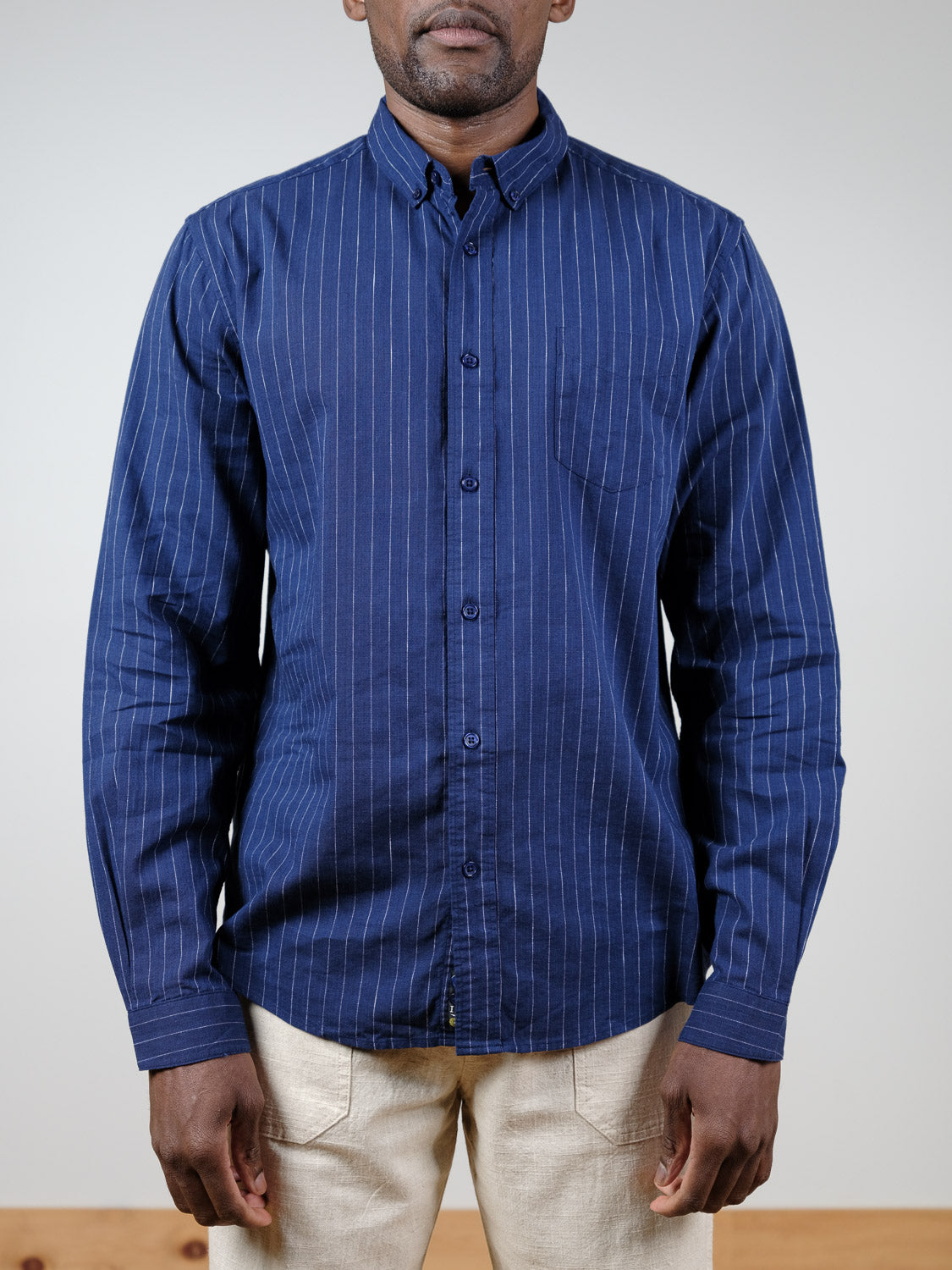 A person, face partially out of frame, stands against a plain light background wearing the Dubai Long Sleeve Shirt by ID Menswear—a blue cotton-linen pinstripe shirt with white vertical stripes—paired with light beige pants.