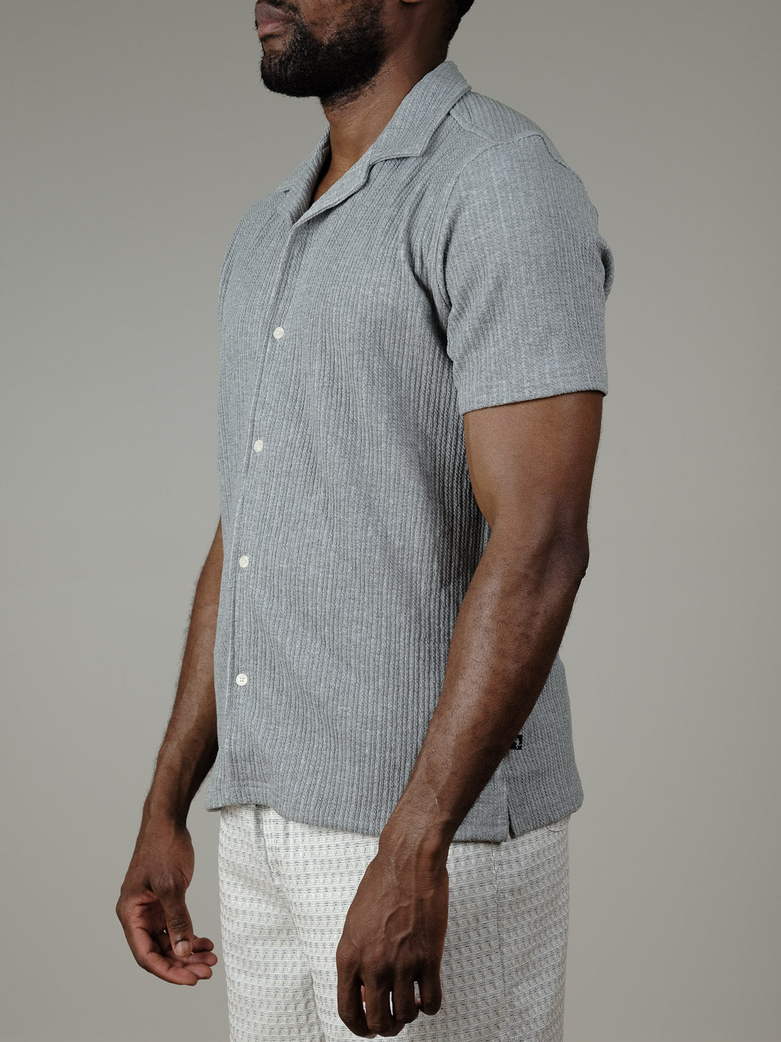 A man stands sideways in a Civil Society Viggo Knit Resort Shirt, light gray with subtle stripes, paired with white patterned shorts. The plain light gray background enhances the shirt’s relaxed vacation vibe.