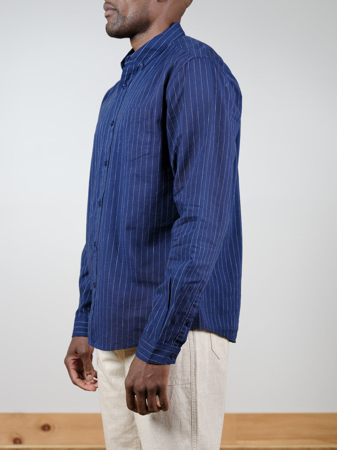 A man stands sideways against a plain background, wearing the Dubai Long Sleeve Shirt by ID Menswear in navy blue pinstripes and a cotton-linen blend, paired with light beige pants.