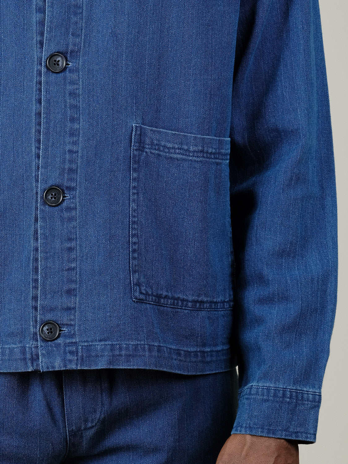 A person is seen wearing the Asplund Herringbone Jacket by Casual Friday, a blue lightweight denim jacket with a front pocket, paired with blue denim pants. Only the lower torso and one arm are visible.