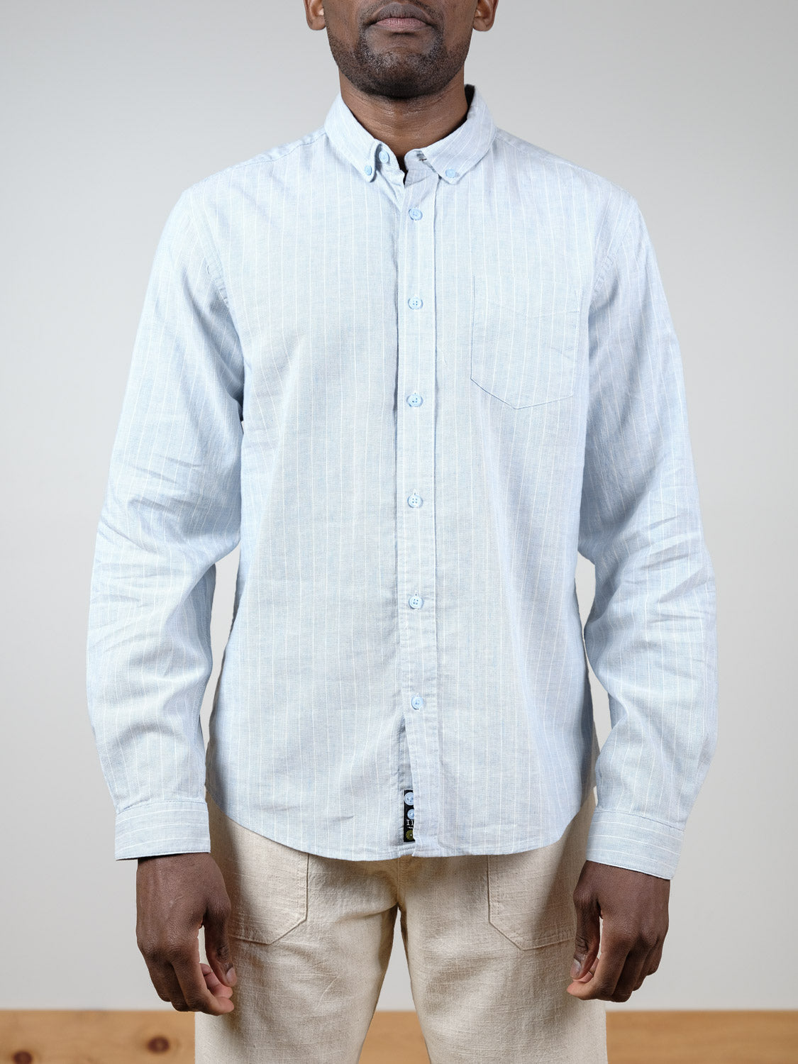 A person wears the Dubai Long Sleeve Shirt by ID Menswear, a light blue cotton-linen pinstripe top, paired with beige pants against a plain light gray background.