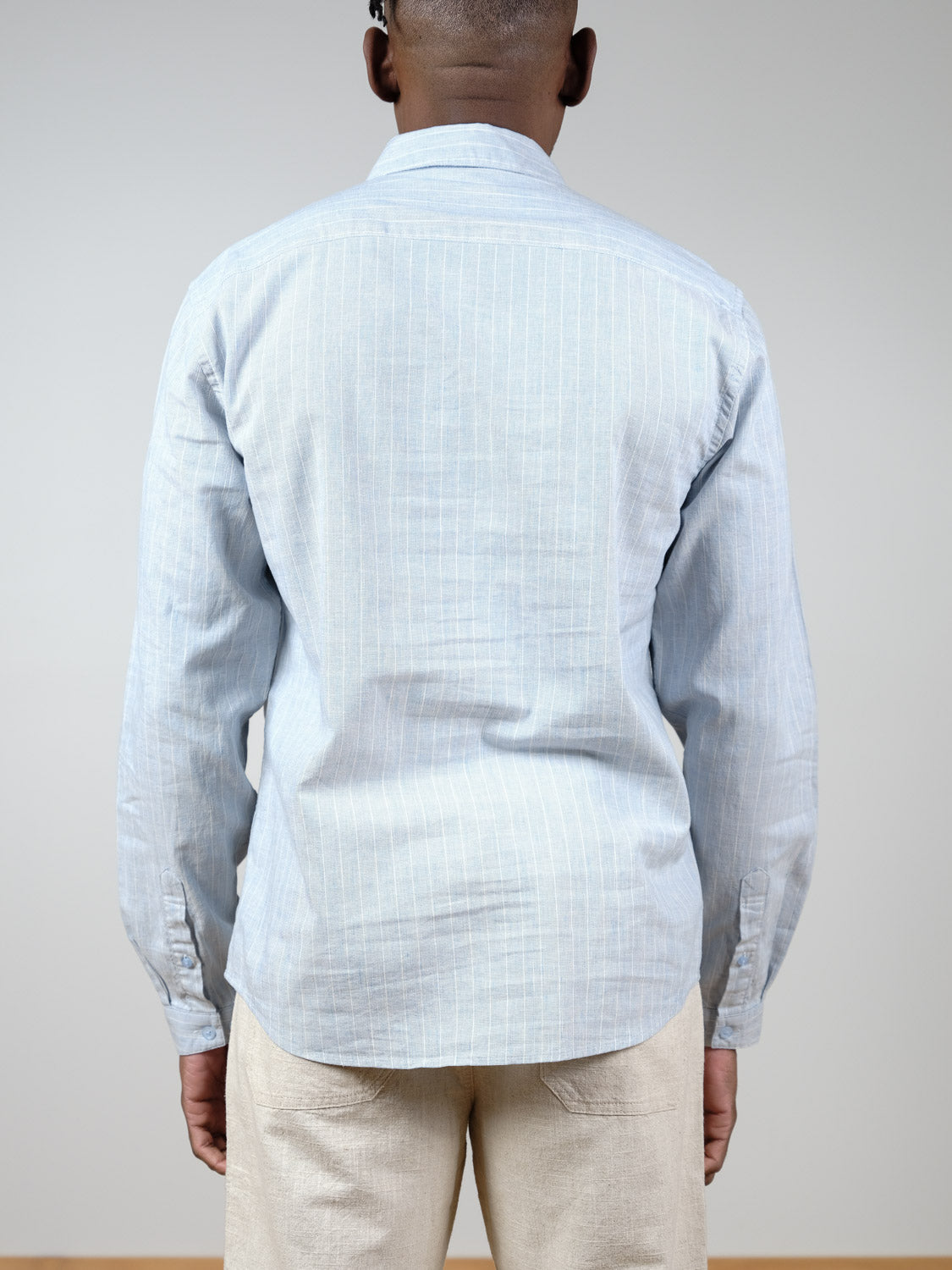 A person faces away from the camera, wearing the Dubai Long Sleeve Shirt by ID Menswear in light blue pinstripes and paired with light beige pants. The background is plain and neutral.