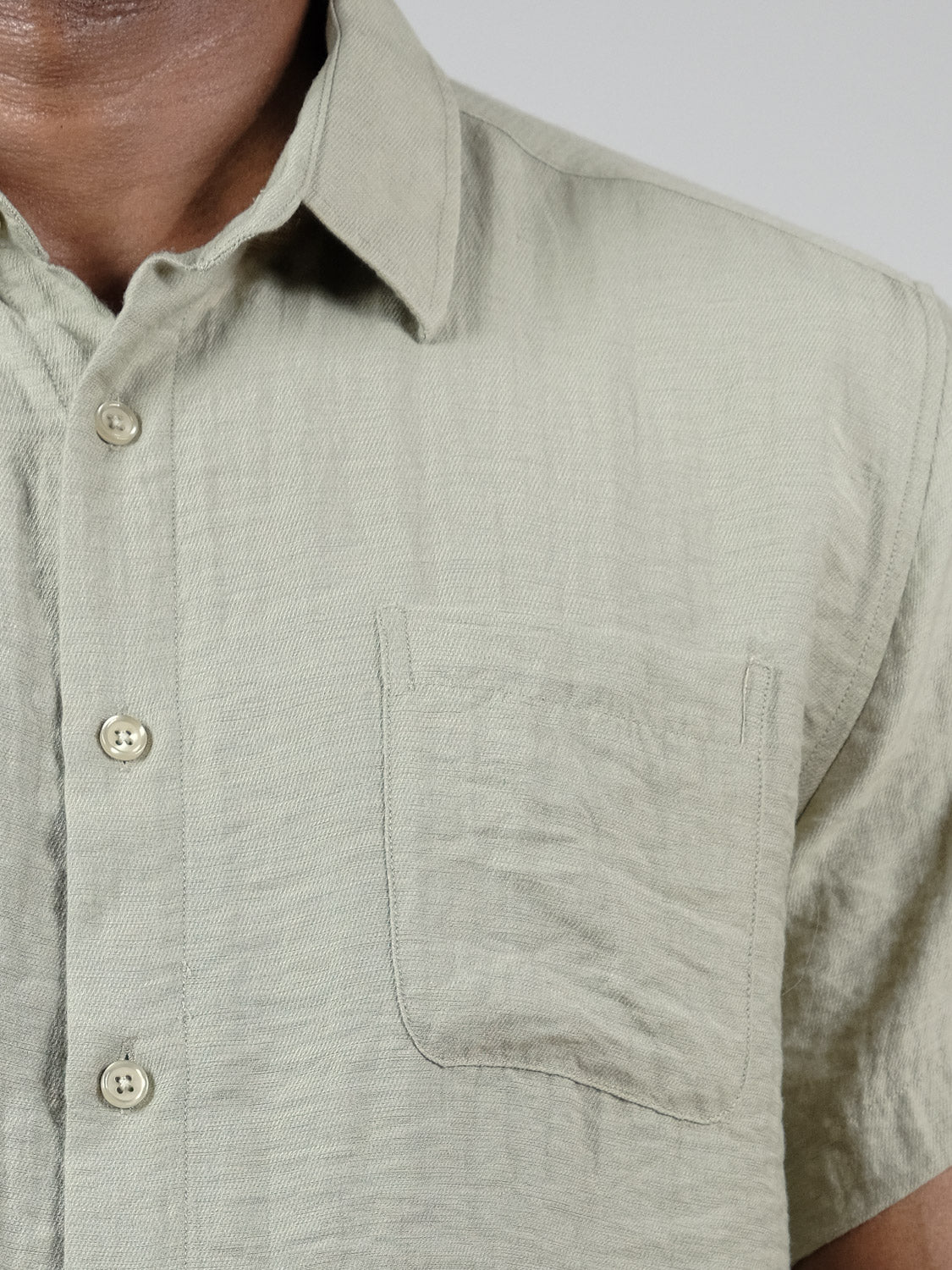 Close-up of a person wearing the ID Menswear Hiker Short Sleeve Shirt in light olive green—a relaxed fit summer essential with a small chest pocket. Only the upper torso is visible.