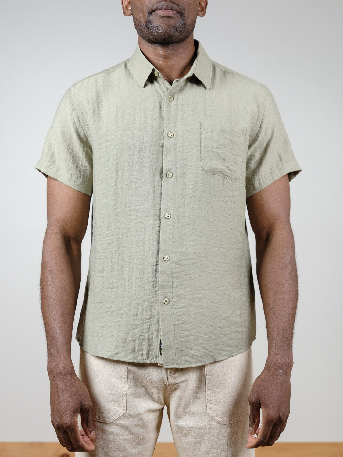 A man models the ID Menswear Hiker Short Sleeve Shirt in light green—a linen button-up with a chest pocket—paired with beige pants. He stands against a plain light background, hands relaxed, exemplifying effortless summer style.