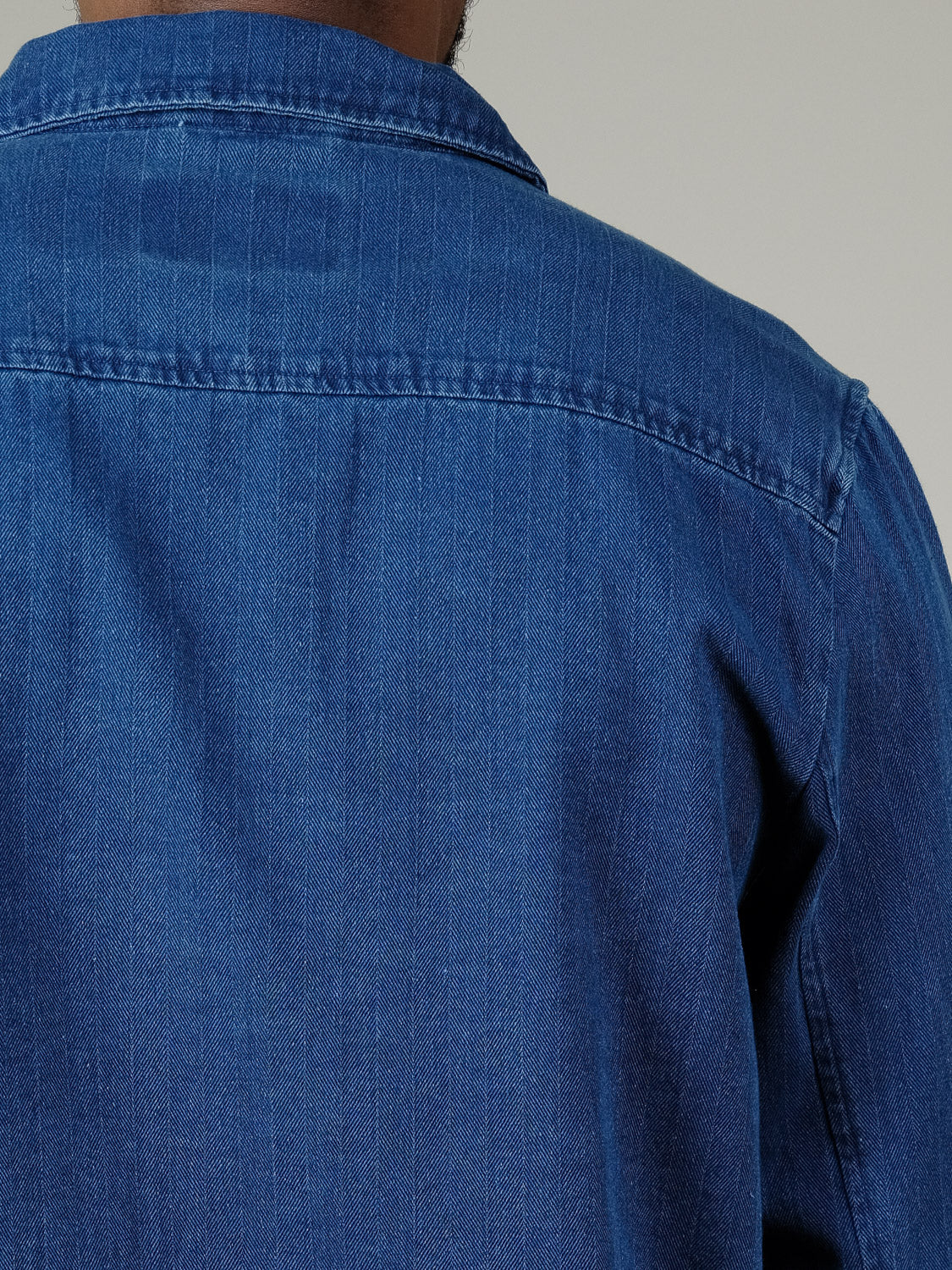 A close-up shows a person from behind wearing the Casual Friday Asplund Herringbone Jacket, highlighting the blue denim fabric, visible stitching, and detailing on the upper back and shoulders.