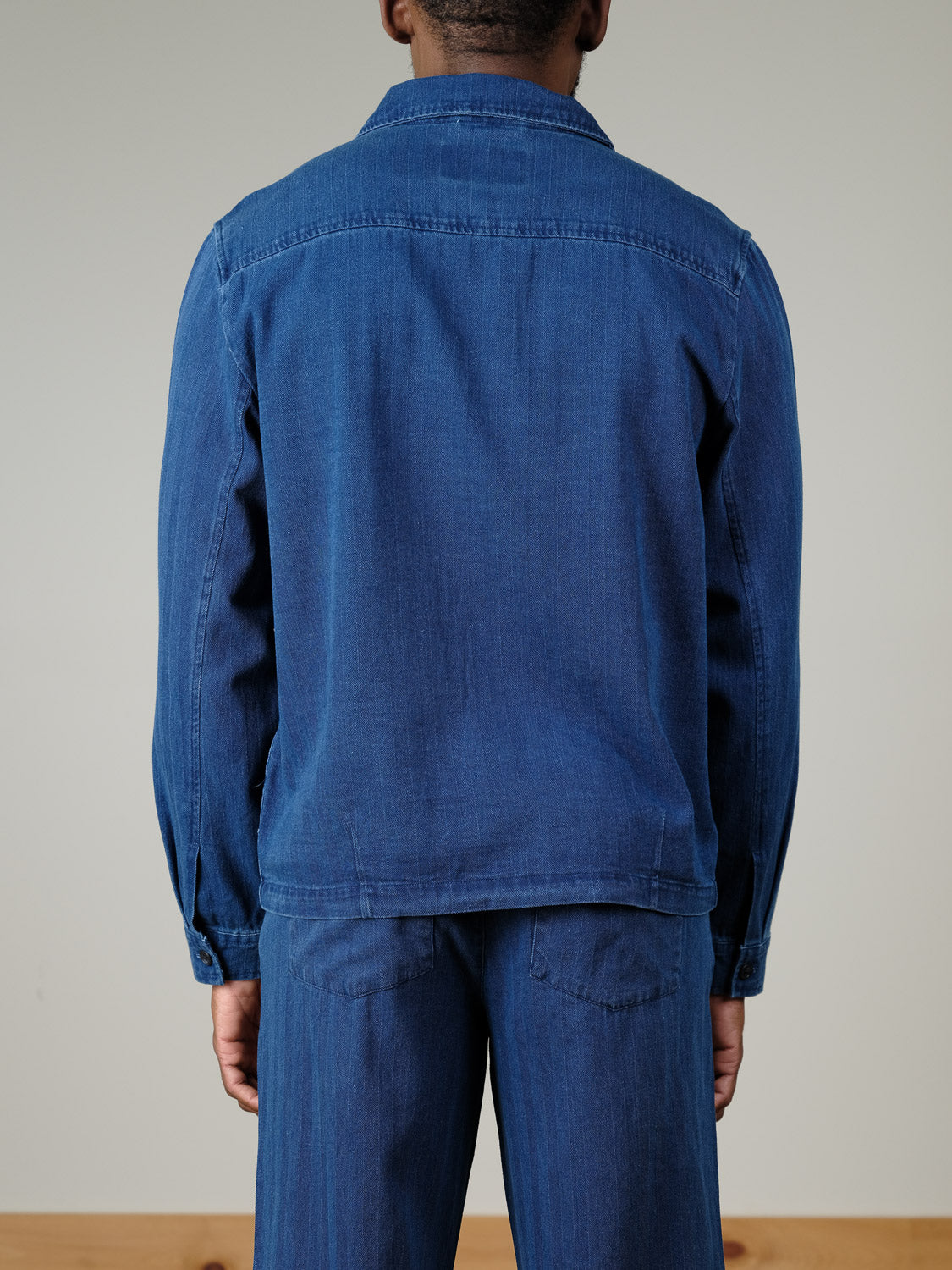 A person stands facing away from the camera, wearing the Casual Friday Asplund Herringbone Jacket in blue with matching pants. The background is plain and neutral.