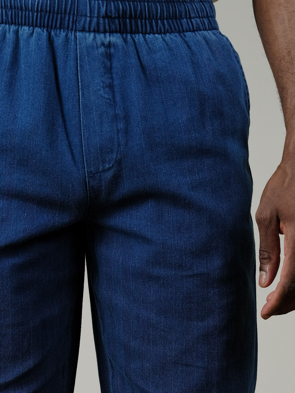 A close-up of a person wearing Casual Friday’s Beck Herringbone Pants, featuring an elastic waistband and durable denim, with their lower torso and right hand visible against a plain background.