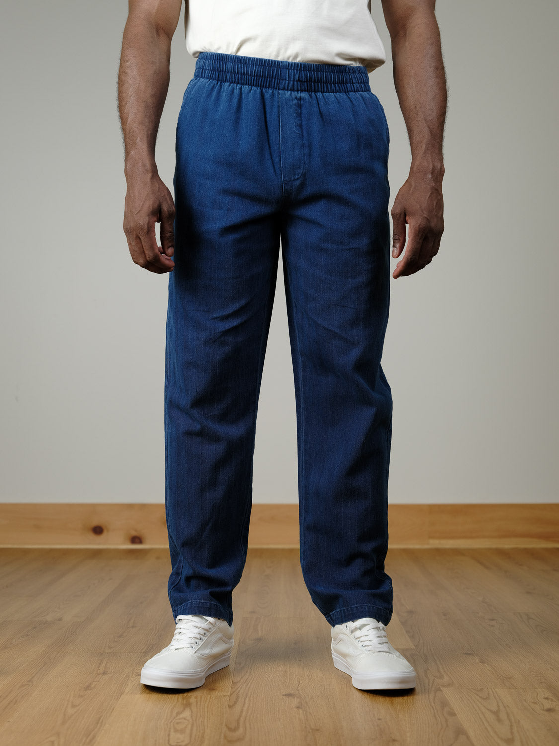 A person wears Casual Friday’s Beck Herringbone Pants in blue, paired with a white T-shirt and sneakers, standing on a wooden floor against a light wall. The chest-cropped photo highlights the relaxed straight-leg elastic-waist pants.