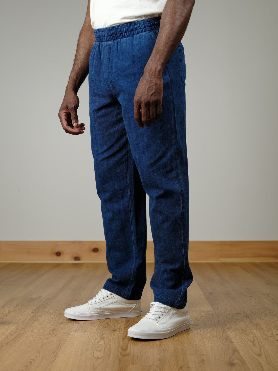 A person stands indoors on a wooden floor, shown from the shoulders down, wearing Casual Friday’s Beck Herringbone Pants in blue with an elastic waistband and white sneakers. The background is plain with light-colored walls and wood trim.