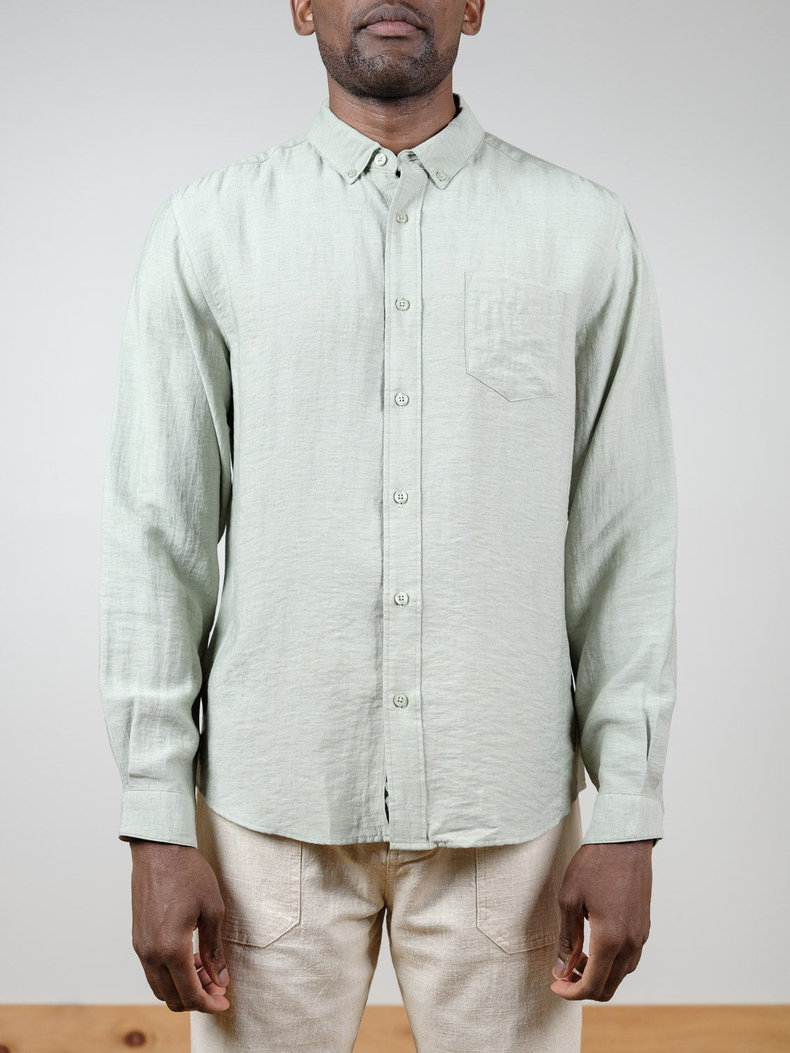 A person faces forward in a relaxed fit, light green Lagos Long Sleeve Shirt by ID Menswear with chest pocket, collar, and long sleeves, paired with beige pants. The breathable linen shirt stands out against a plain neutral background.