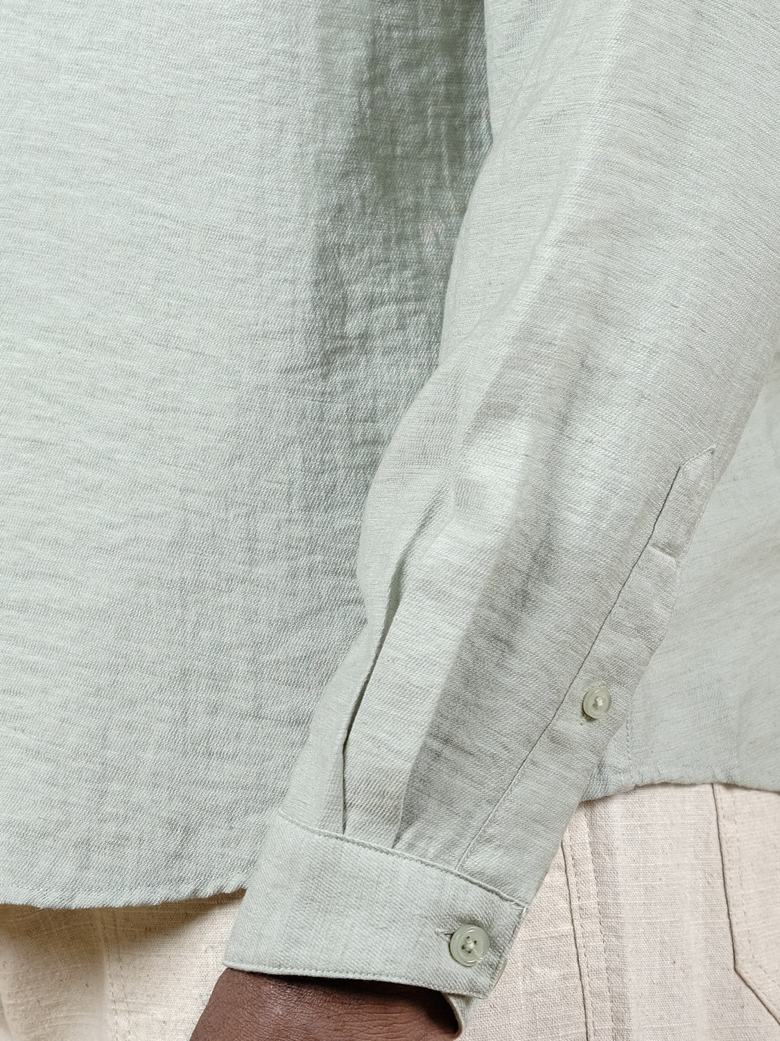 A close-up shows a person wearing the Lagos Long Sleeve Shirt by ID Menswear in light green linen, paired with cream pants. Only the arm and torso are visible, highlighting its relaxed fit and textured, breathable fabric.