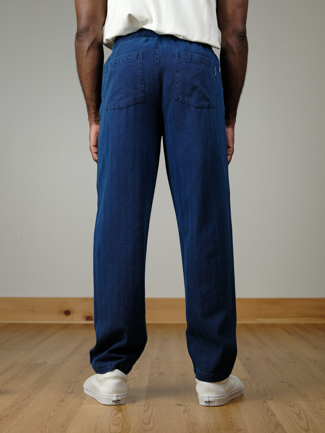 A person stands indoors on a wooden floor, seen from the back, wearing a white shirt, Casual Friday Beck Herringbone Pants in blue with back pockets, and white sneakers.