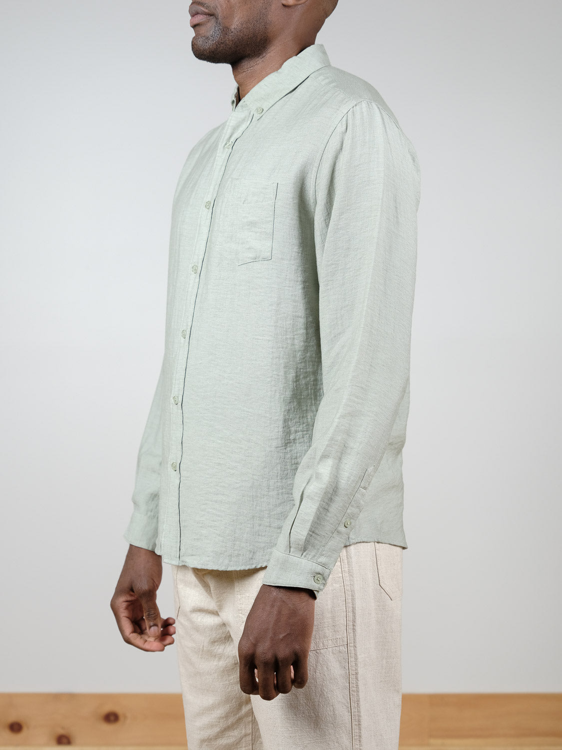 A person stands in profile against a plain background wearing ID Menswear's Lagos Long Sleeve Shirt in light green linen with a chest pocket, paired with beige pants.