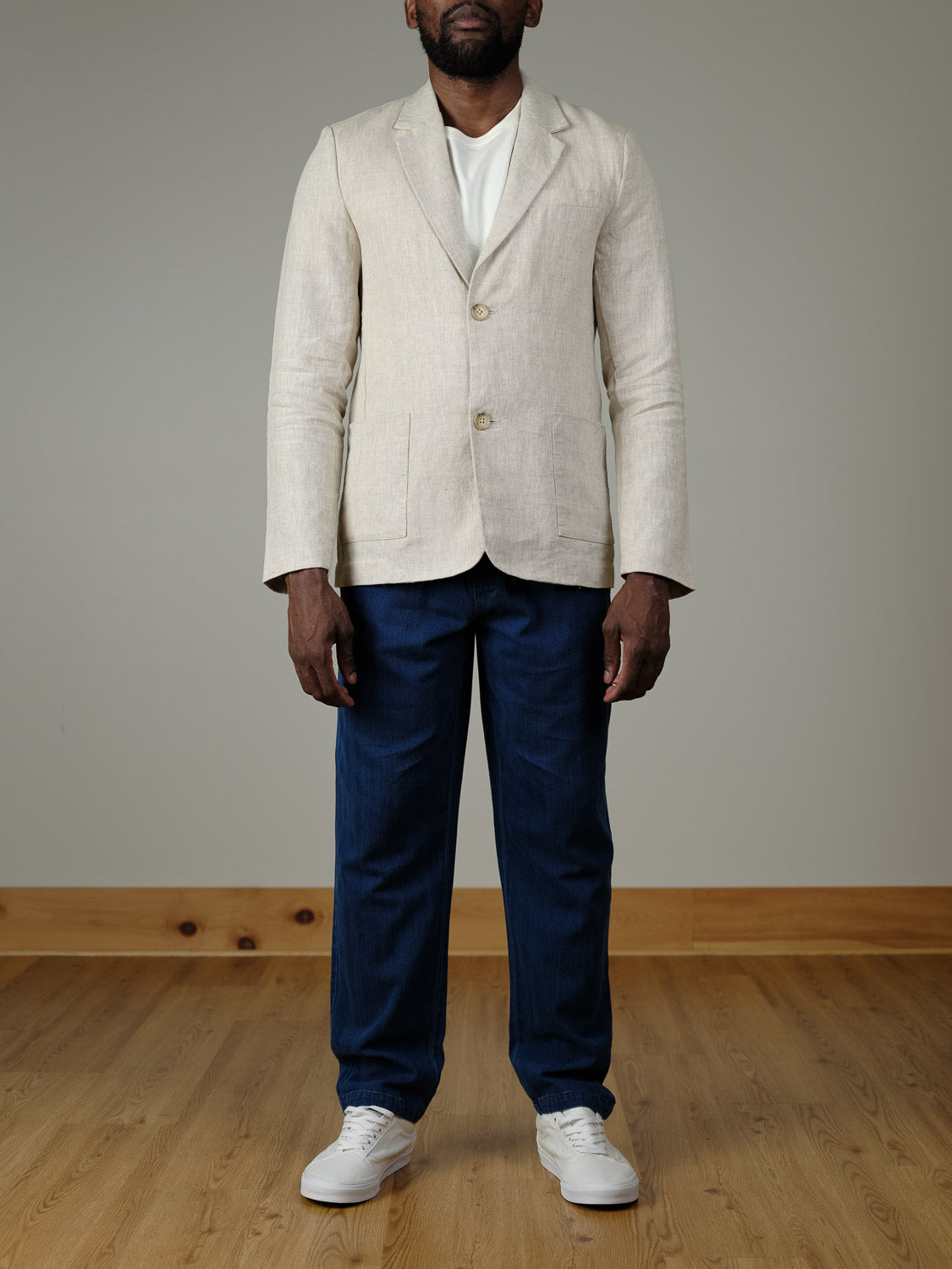 A man wears the Casual Friday Torup Linen Blazer in light beige over a white shirt, with dark blue pants and white sneakers. He stands indoors against a plain light wall and wooden floor, his face partially out of frame.