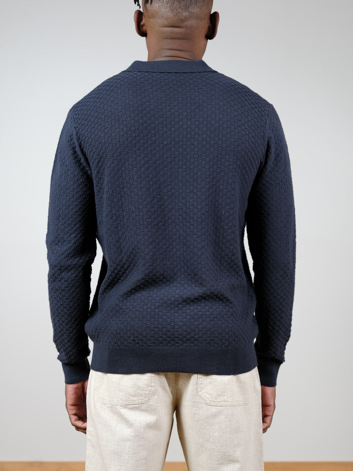 A person faces away from the camera, wearing an ID Menswear Luxor Long Sleeve Cotton Polo and light beige tailored pants, set against a plain, light background.