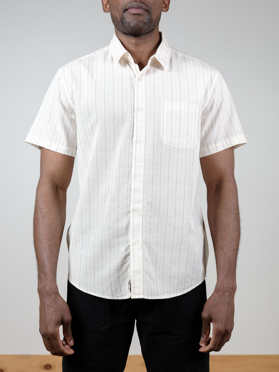 A man wears the Tripper Short Sleeve Shirt by ID Menswear, featuring thin vertical stripes, a chest pocket, and a light color, standing against a plain background.
