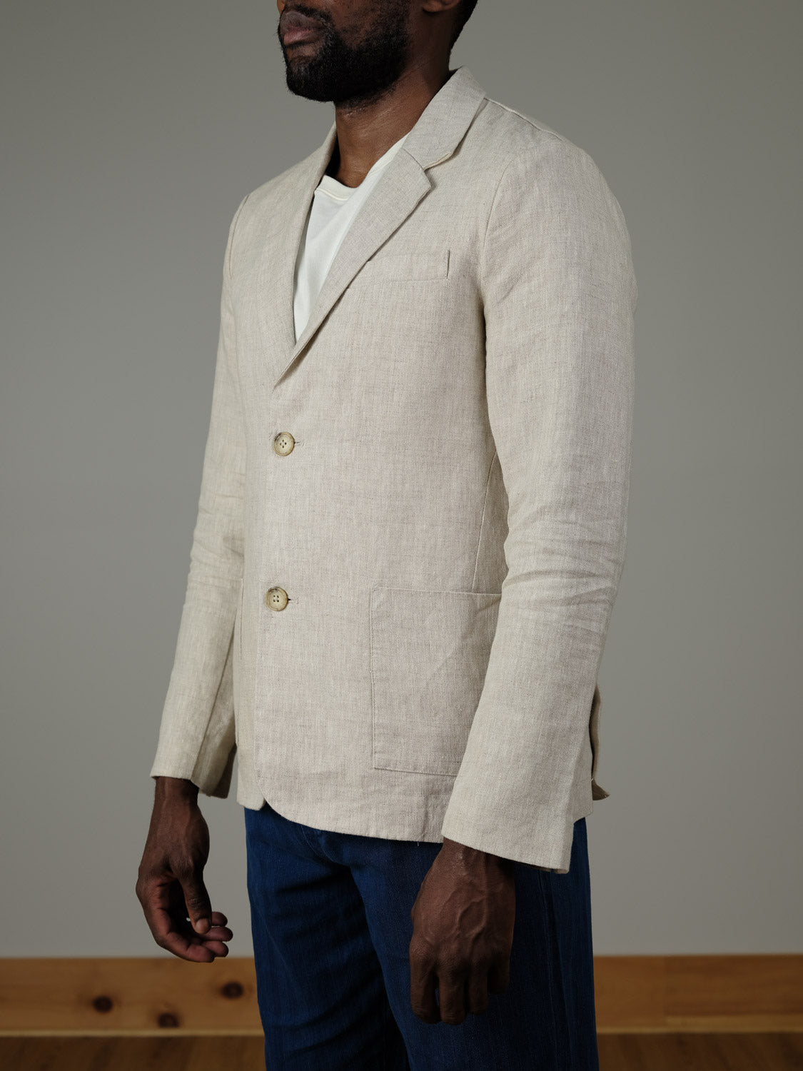 A man models the Torup Linen Blazer by Casual Friday—a light beige, unstructured jacket—over a white shirt and dark blue jeans, standing against a plain background for an effortlessly stylish summer look.