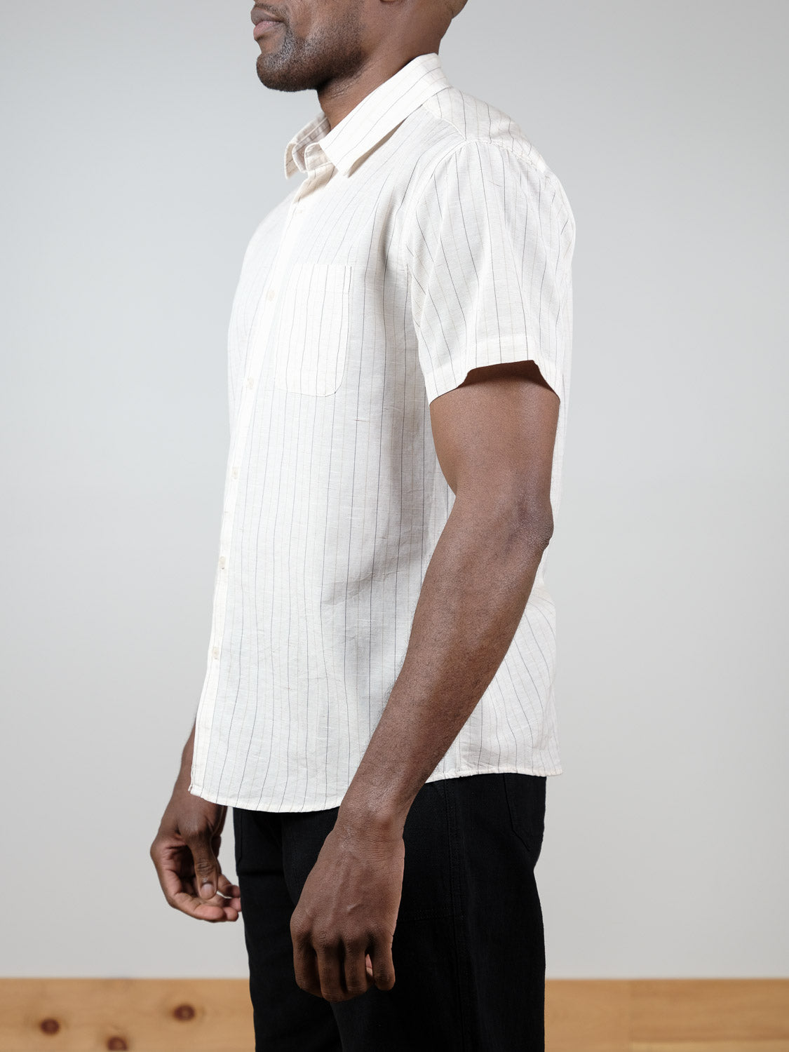 A man stands sideways in a Tripper Short Sleeve Shirt by ID Menswear, featuring subtle vertical stripes, paired with black pants against a plain background.