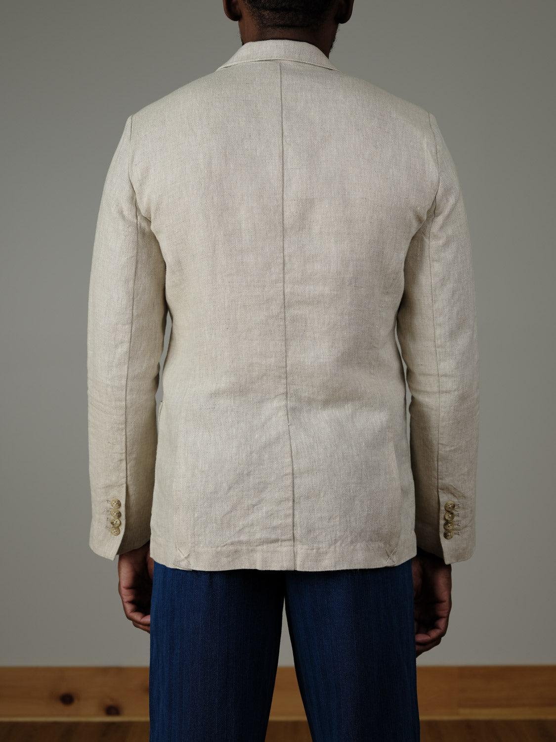 A person stands facing away from the camera, wearing the Casual Friday Torup Linen Blazer in light beige and dark blue pants, against a plain indoor background with a light gray wall and wooden floor trim.