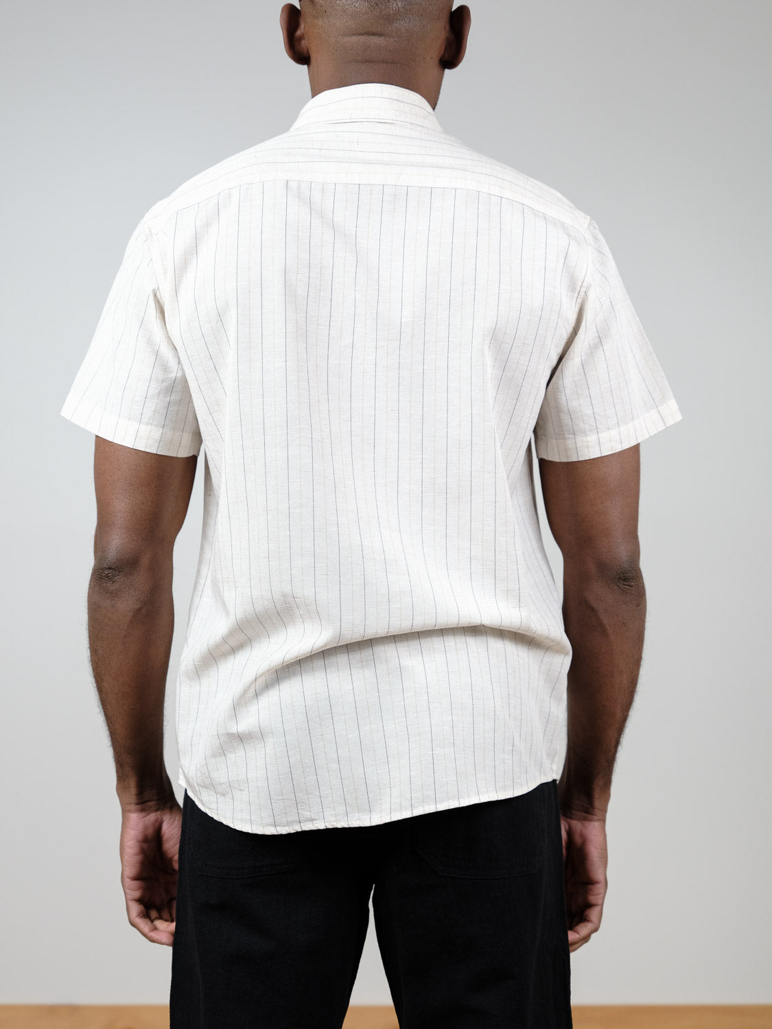 A person stands with their back to the camera, wearing the ID Menswear Tripper Short Sleeve Shirt with light-colored vertical stripes and black pants against a plain background.