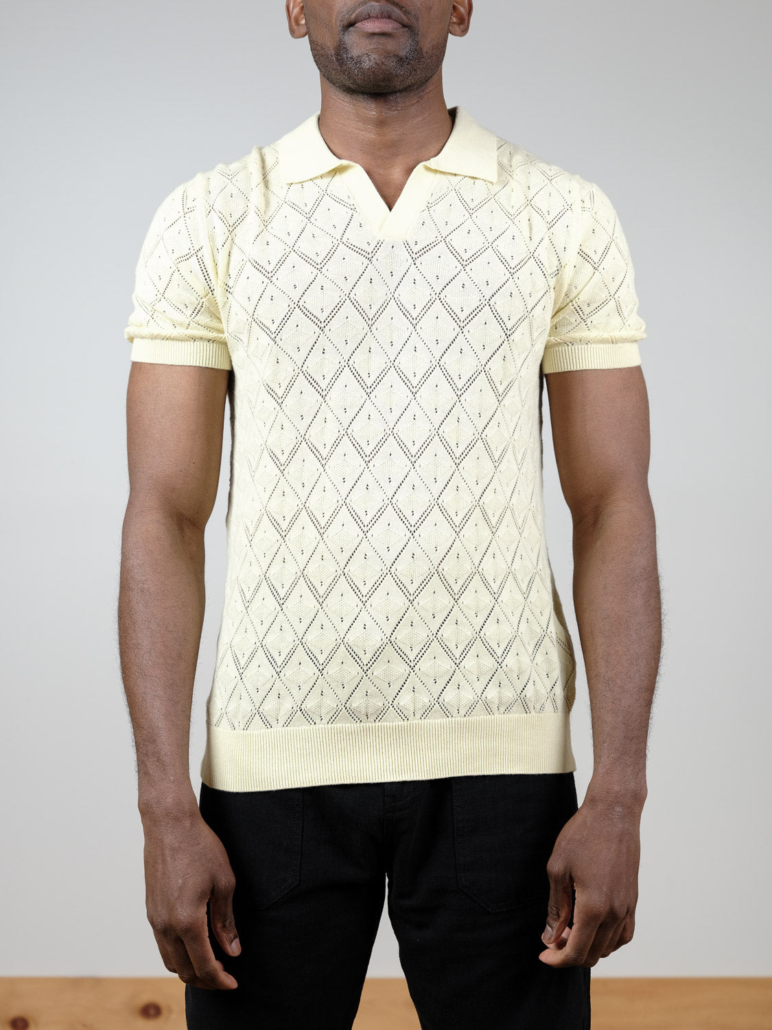 A man wears the Agra Knitted Cotton Polo by ID Menswear, featuring a light yellow color, short sleeves, and a diamond pattern—perfect for casual wear. He stands facing forward against a plain background with black pants.