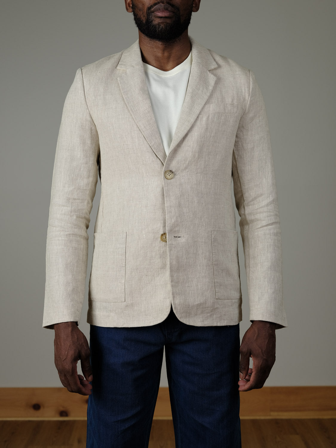 A person stands facing forward against a plain, light background, wearing the Casual Friday Torup Linen Blazer in light beige over a white t-shirt and dark blue pants.