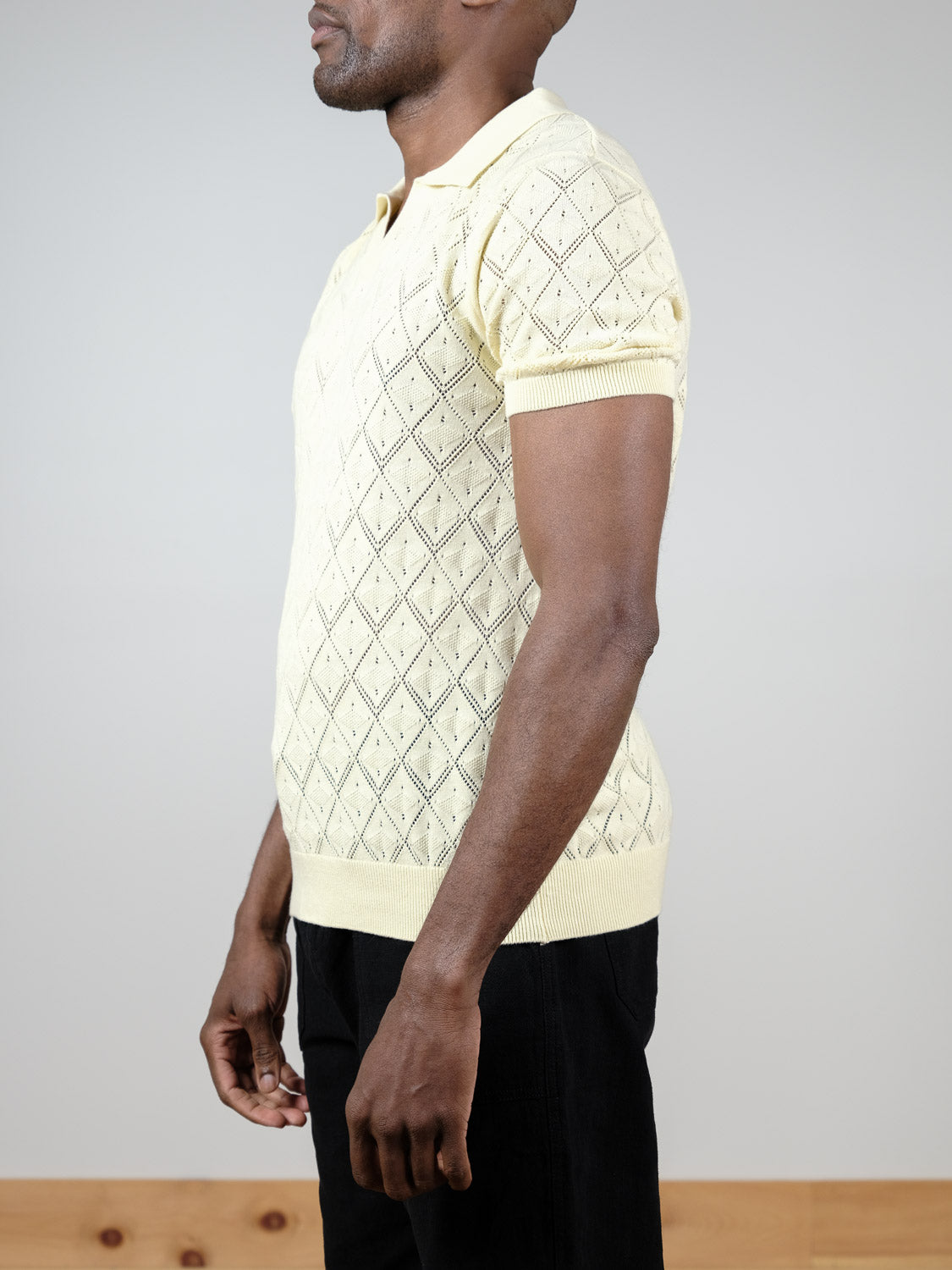 A man stands sideways against a plain background wearing the Agra Knitted Cotton Polo by ID Menswear—a pale yellow, short-sleeved, argyle-patterned cotton shirt—paired with black pants. His face is partially obscured.