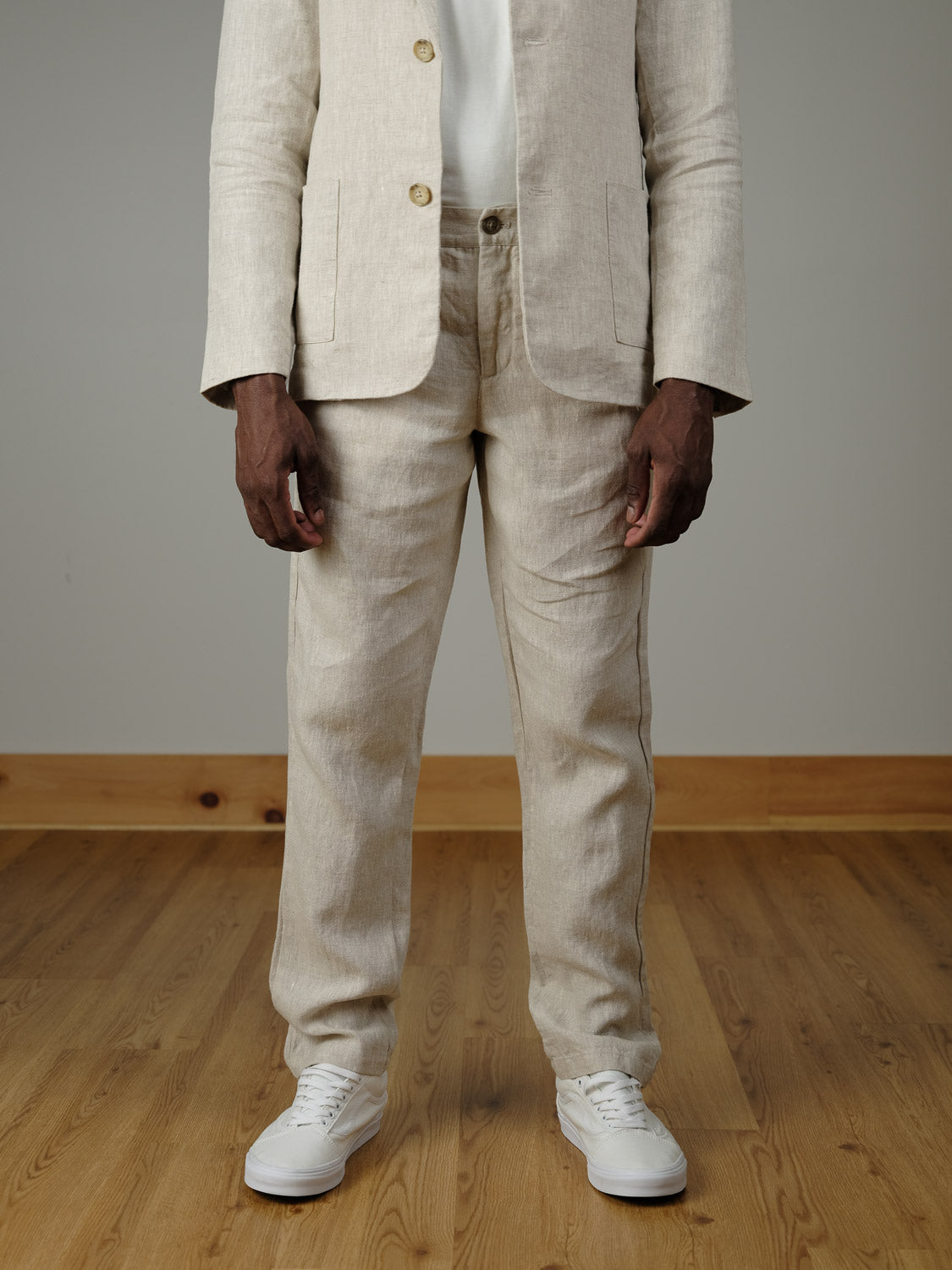 A person wearing Pandrup Linen Pants by Casual Friday—light beige pants with an elastic waistband and white sneakers—stands on a wooden floor, shown from shoulders down, arms relaxed at their sides against a plain background.